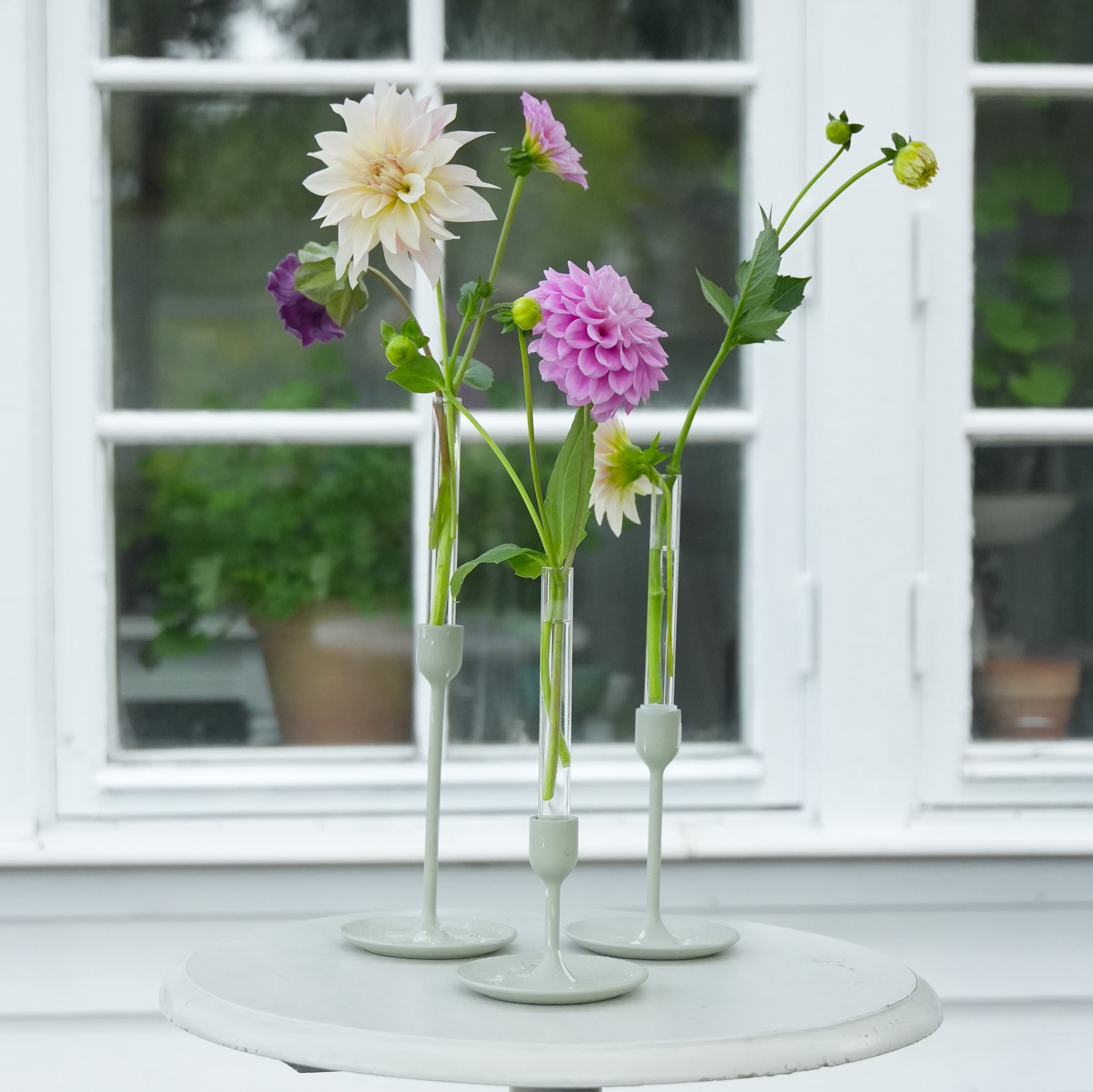 Three floral candle vase holders with Dahlias and green flowers on a white table in front of a window.