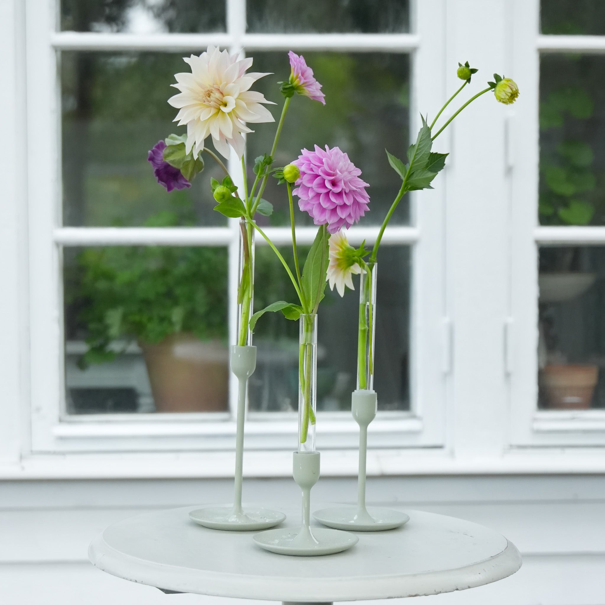 Three floral candle vase holders with Dahlias and green flowers on a white table in front of a window.
