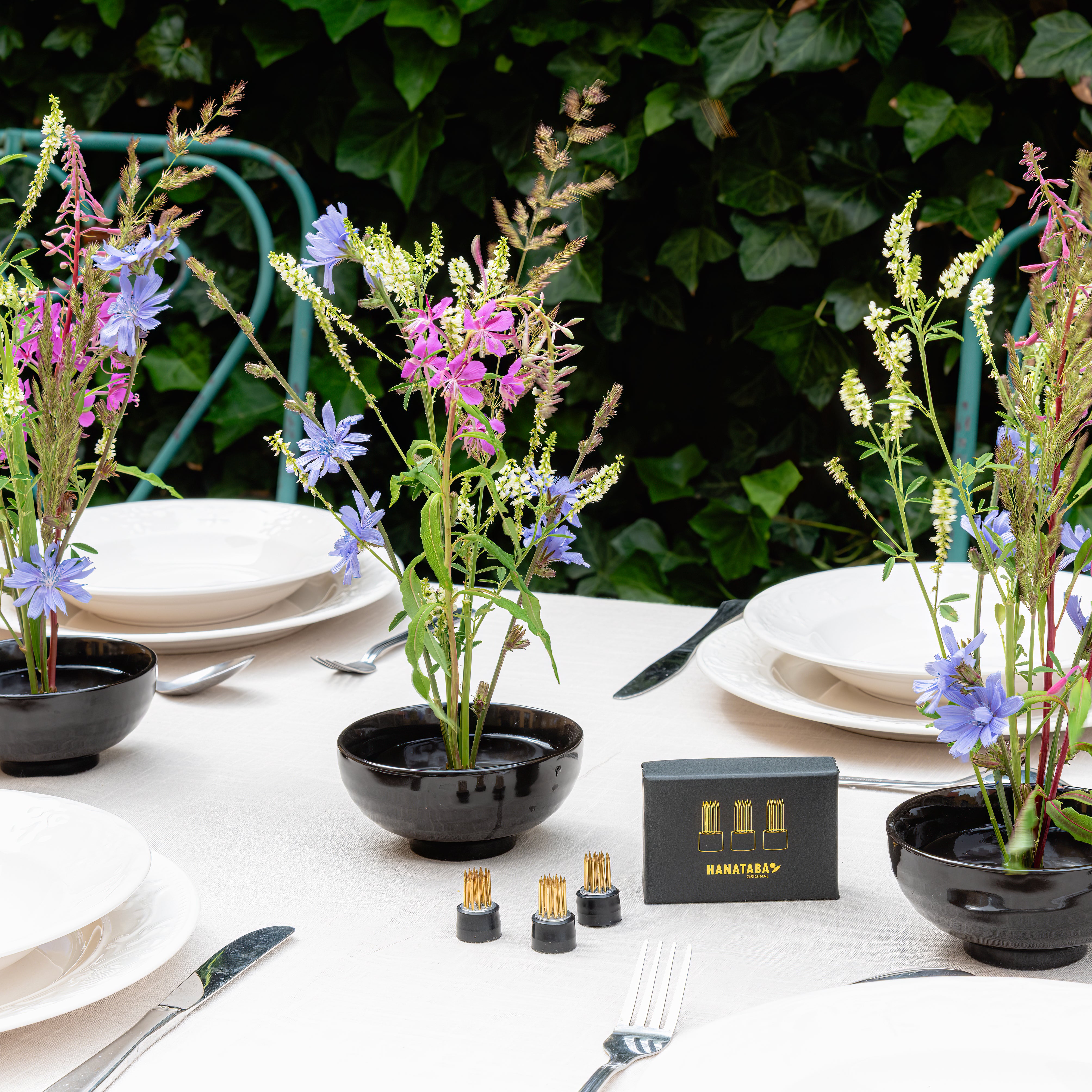 Table setting with black ceramic kenzan bowls containing wildflowers, plates, and cutlery against a green outdoor background.