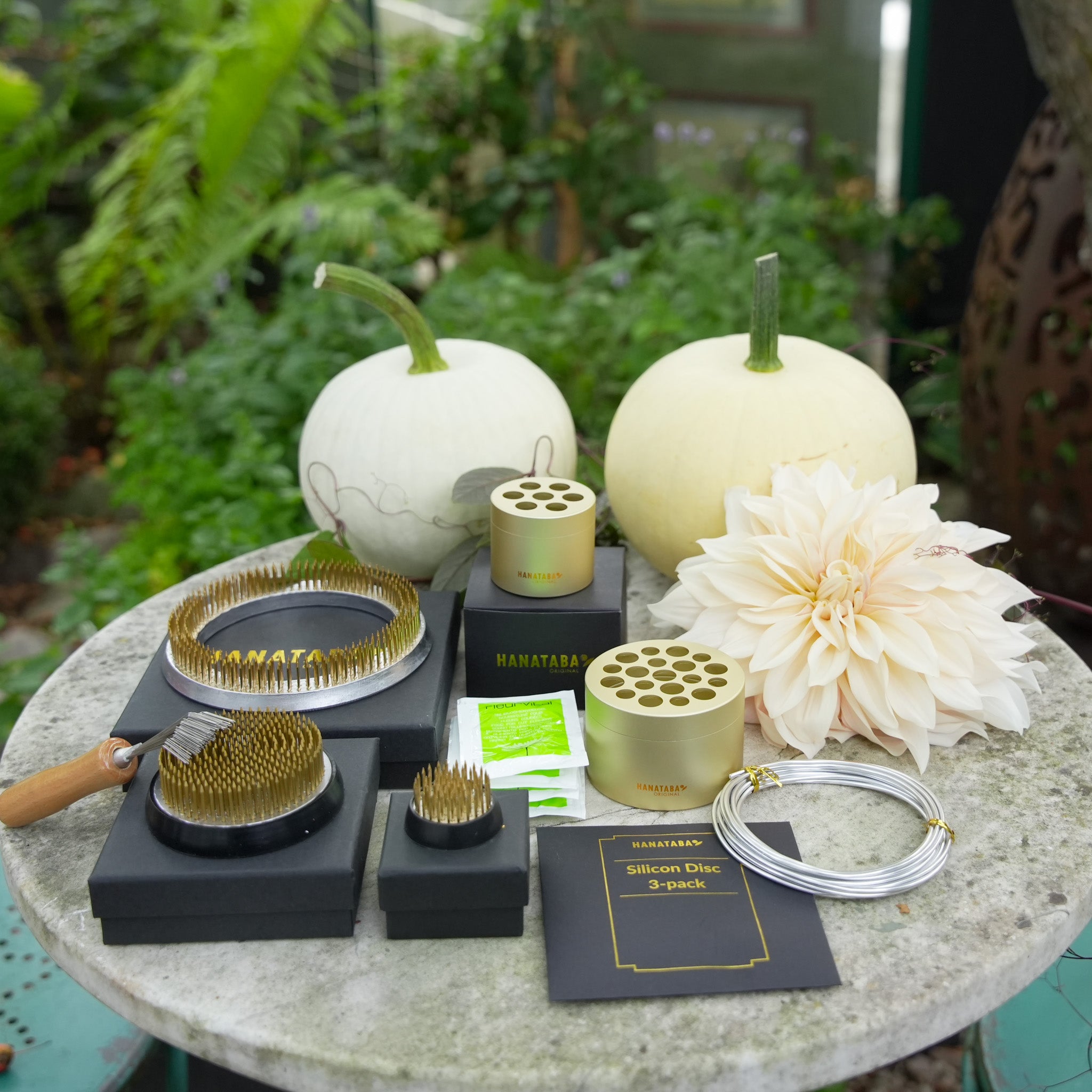 Table with two Hanataba Champagne Gold, 5x flower food, pink bonsai wire, Kenzan flower frog ring 120 mm, kenzan flower frog 70mm and kenzan flower frog 34 mm beside a large white dahlia and two white pumpkins