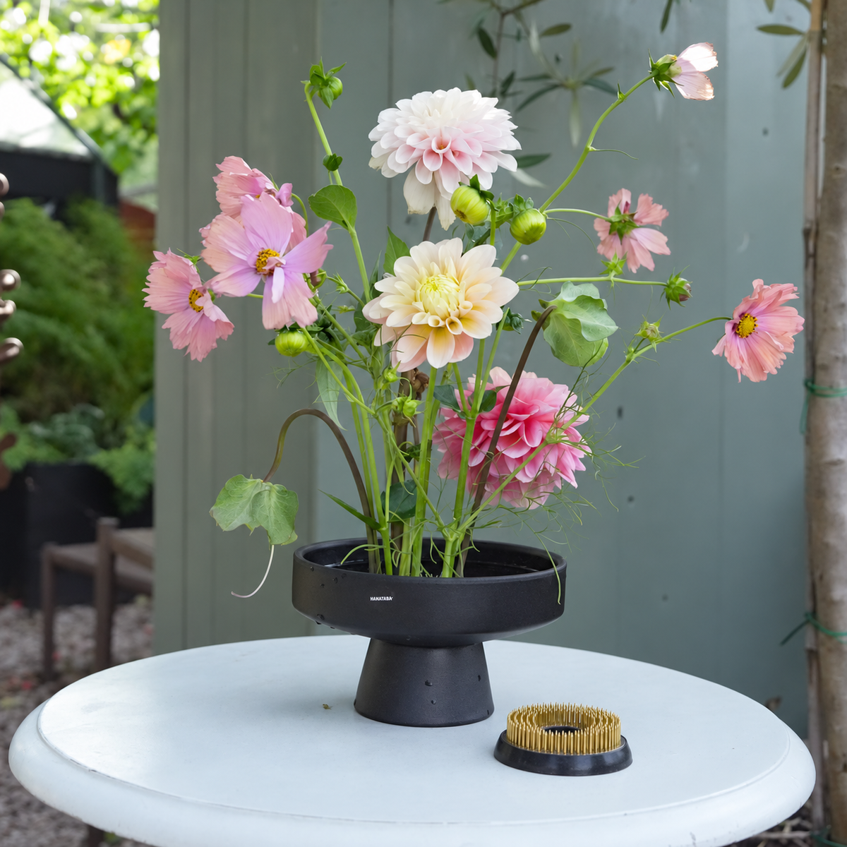 A black floral stand holding pink and cream dahlias next to a Hanataba Original Kenzan ring 70 mm, set on a white round table outdoors with a green fence in the background.