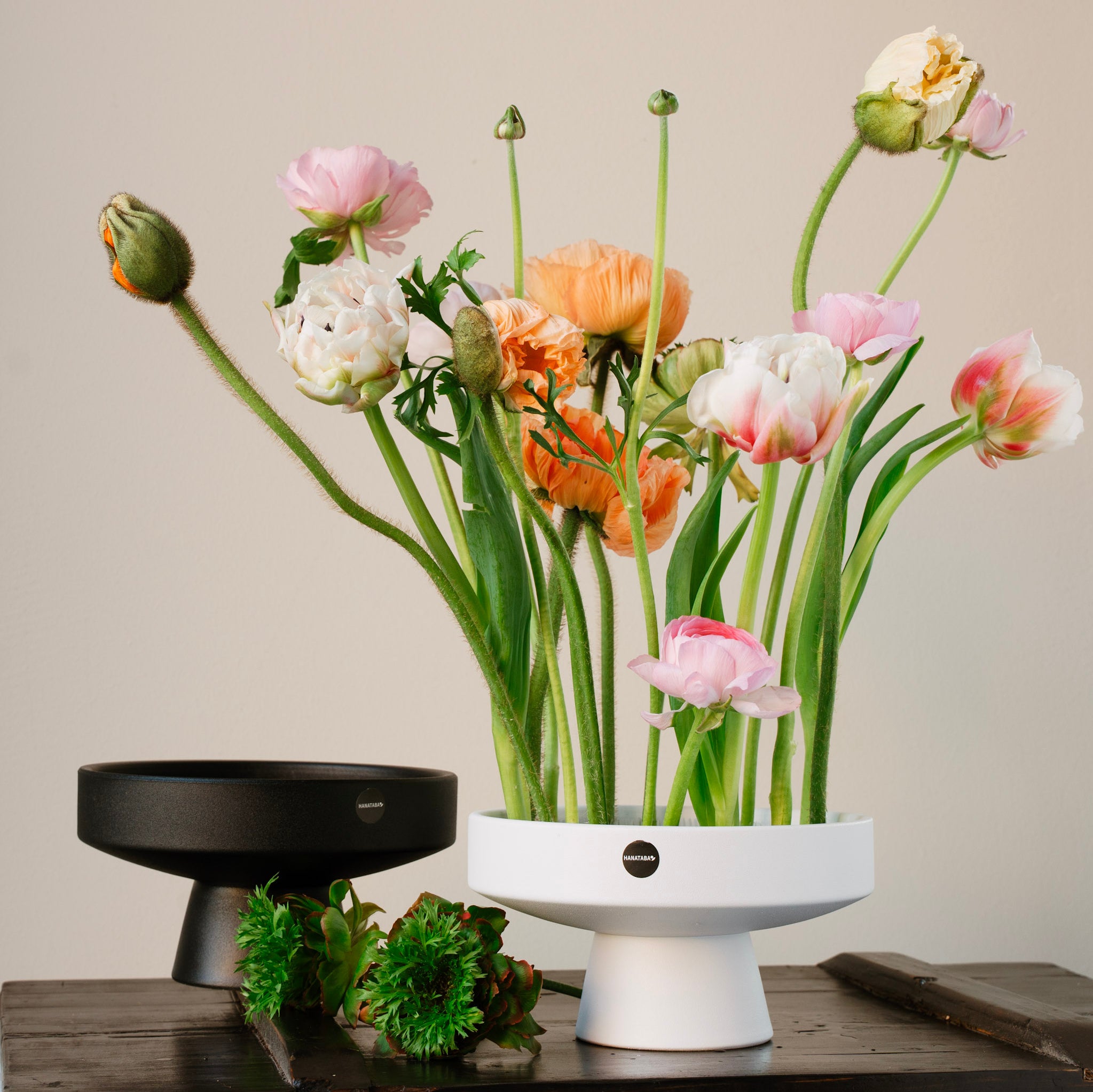 Floral arrangement in a white hanataba ceramic ikebana vase on a wooden surface with a neutral background and a black hanataba ceramic ikebana vase in the background