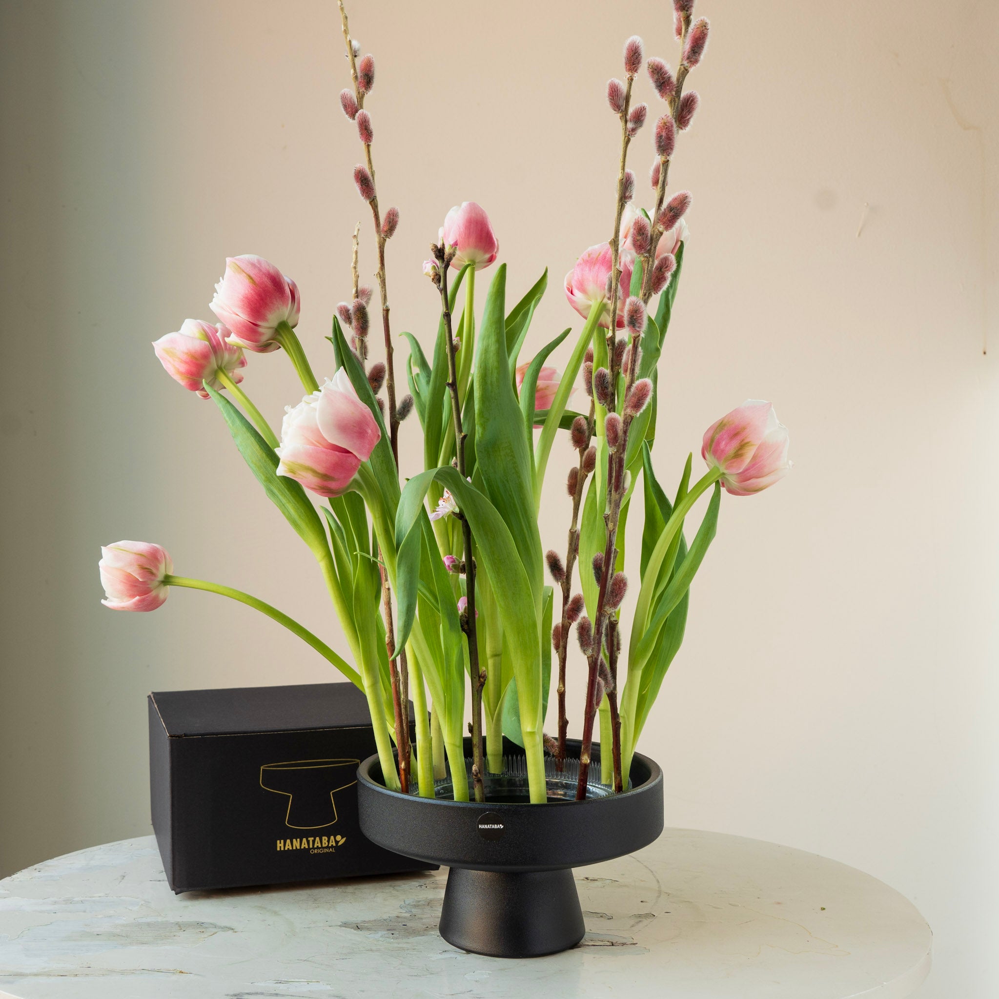 Floral arrangement with pink tulips and willow in a black hanataba ceramic ikebana vase on a light surface.