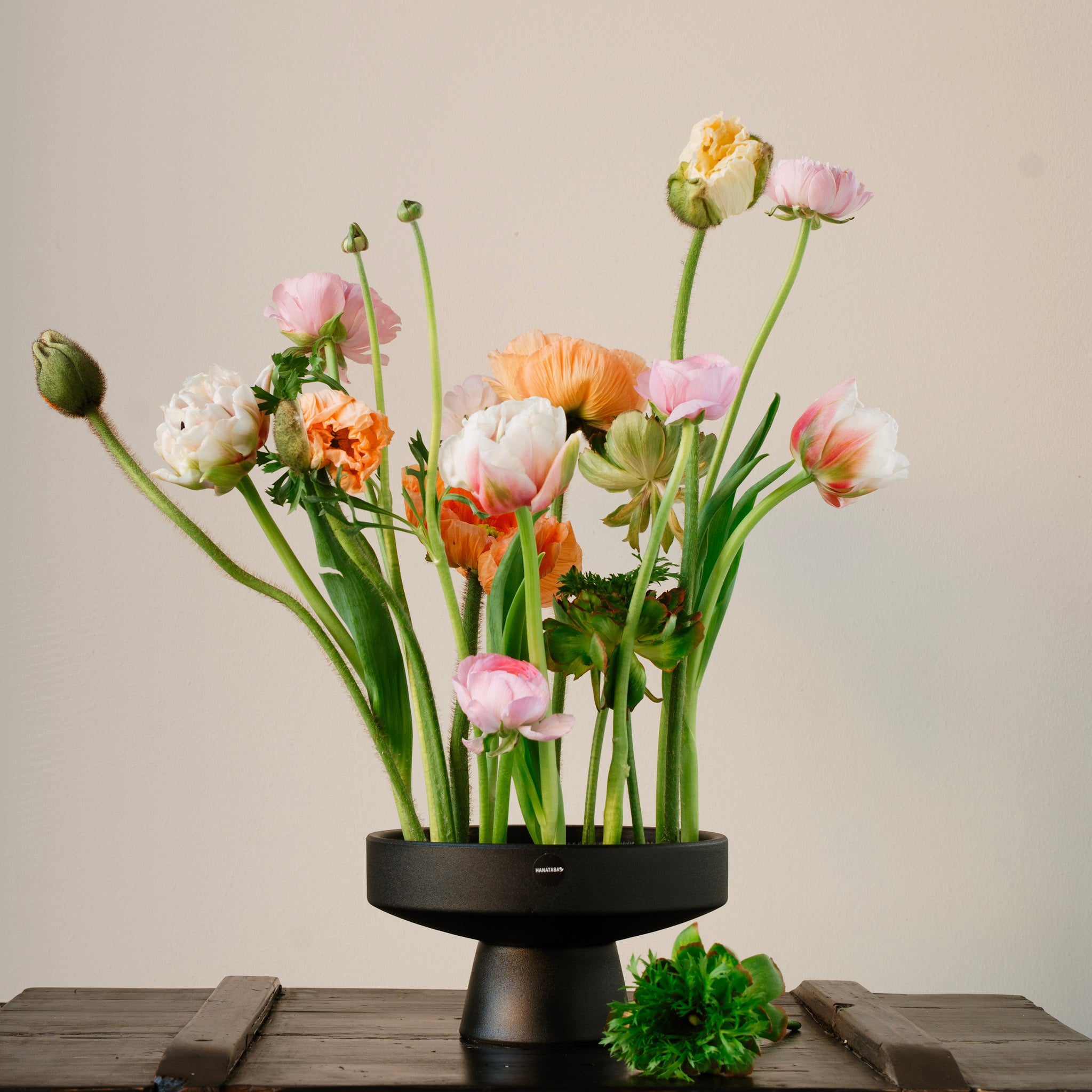 Floral arrangement in a black hanataba ceramicikebana vase on a wooden surface with a neutral background