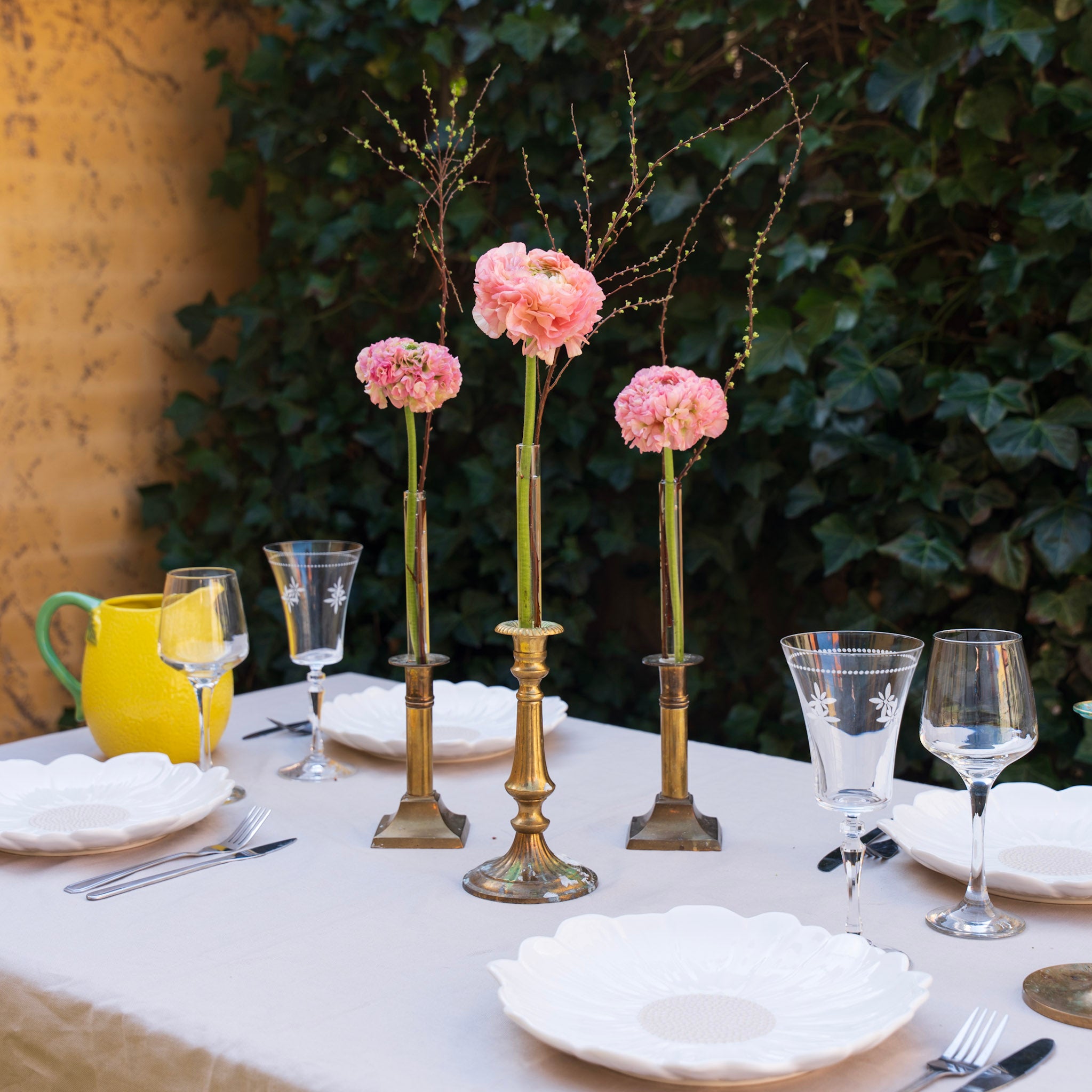 Decorative table setting with pink flowers inside hanataba Candle vase, glasses, and cutlery against a green leafy background.