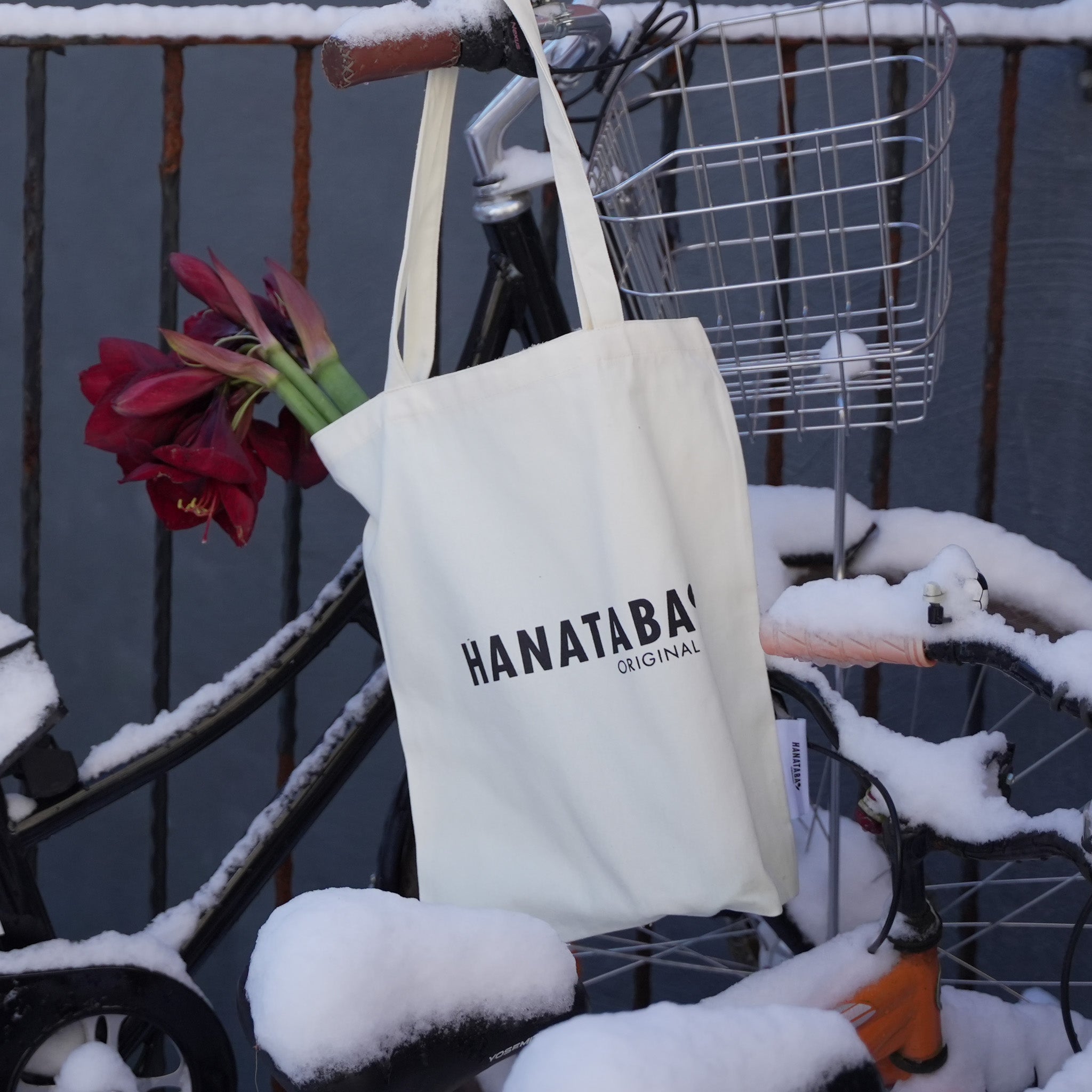 A white Hanataba Original tote bag hanging on a snow-covered bicycle with red amaryllis flowers peeking out of the bag.