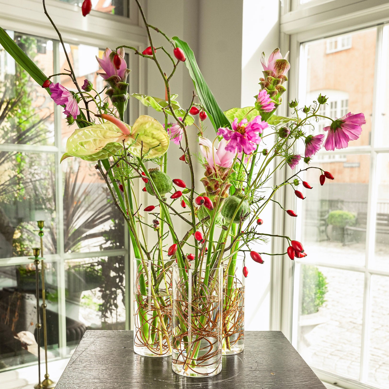 Decorative floral arrangement using brown bonsaiwire in clear vases on a table with large windows in the background.