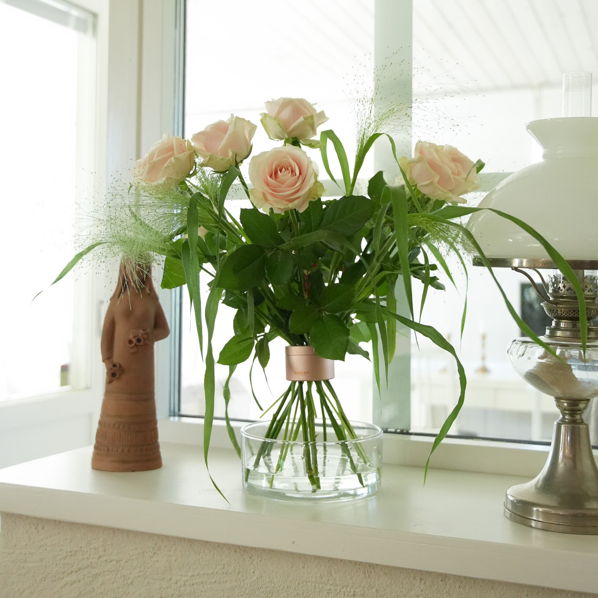 Bouquet of pink roses in a hanataba pink champagne. Standing on a clear vase on a windowsill with a lamp and decorative object in the background.