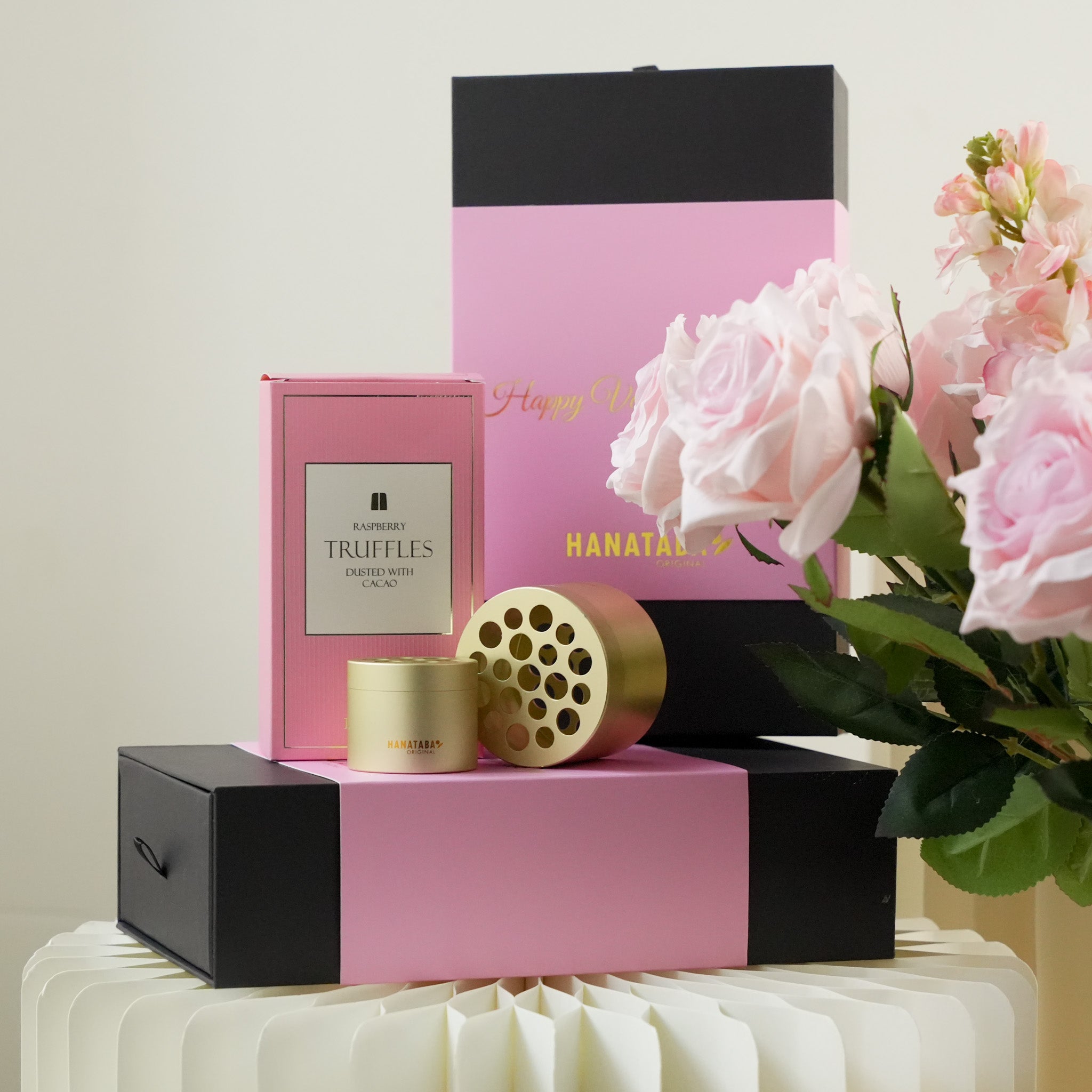 Pink and black gift box with a gold cylindrical object called Hanataba Bouquet twister and a bouquet of pink roses on a white surface.