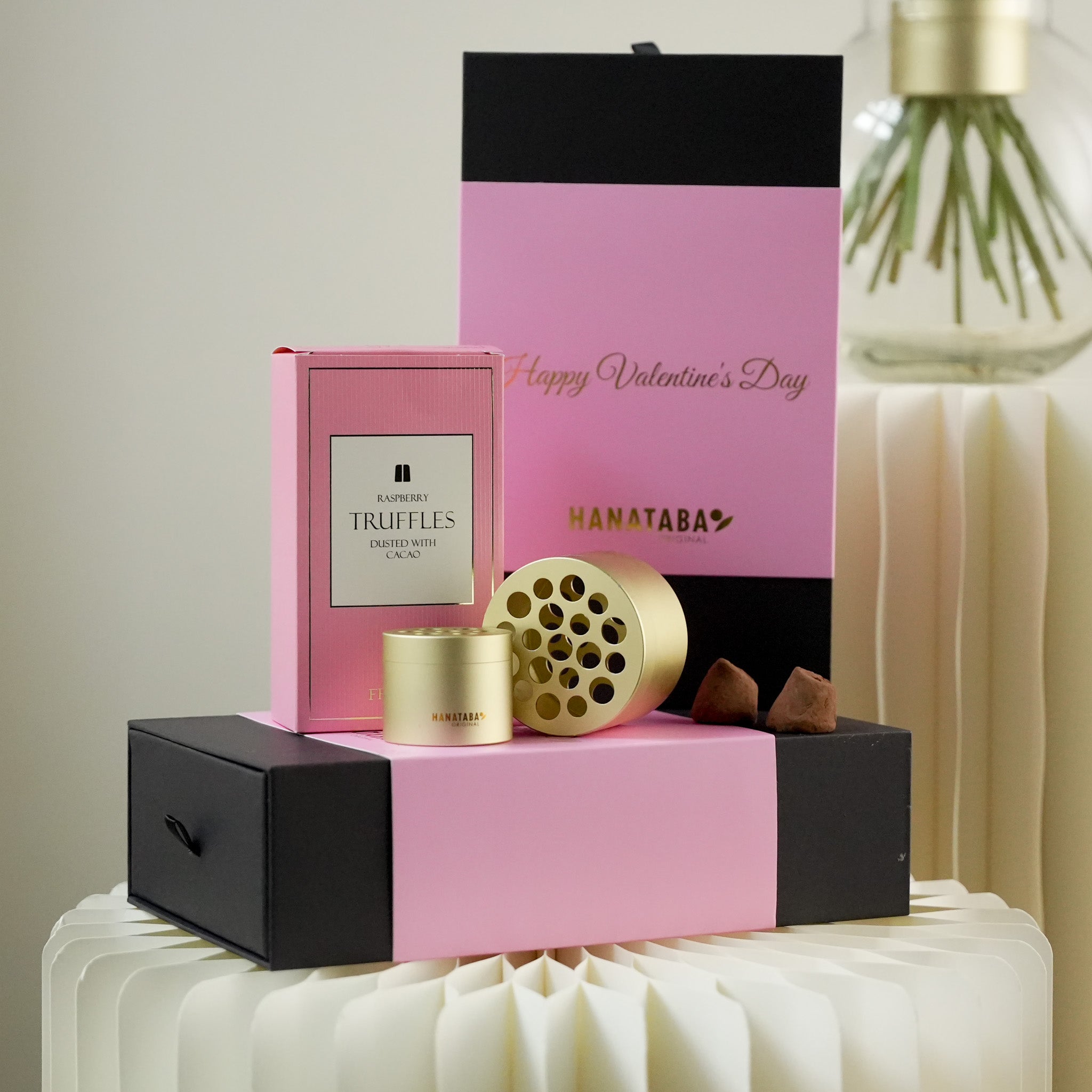 Valentine's Day gift set with pink and black box, truffles, and a gold flower tool (Hanataba) on a white surface.