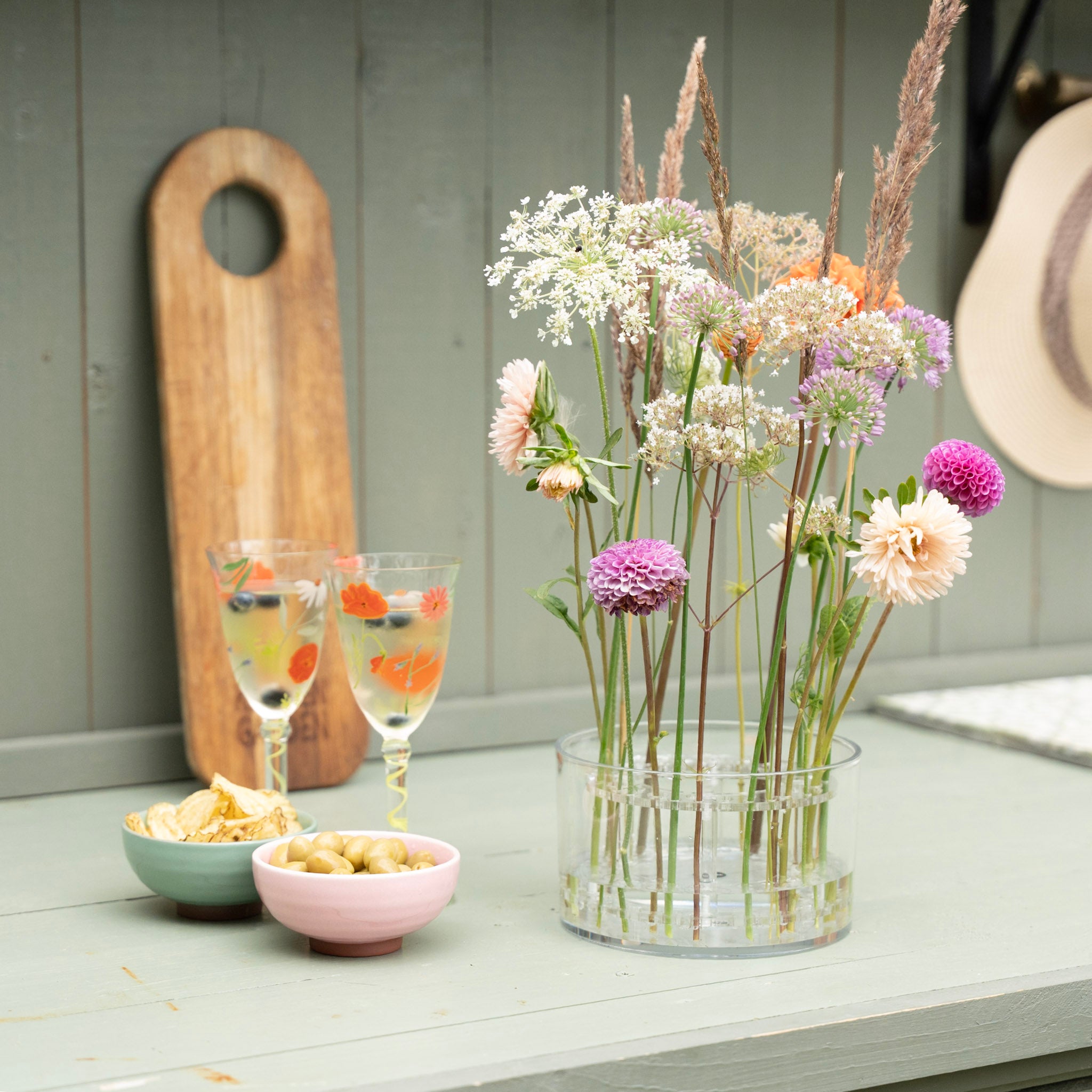 A clear glass vase filled with a bouquet of wildflowers, including white umbellifers, purple alliums, and peach dahlias, displayed on a light green wooden table next to two glasses of iced tea and small bowls of snacks.