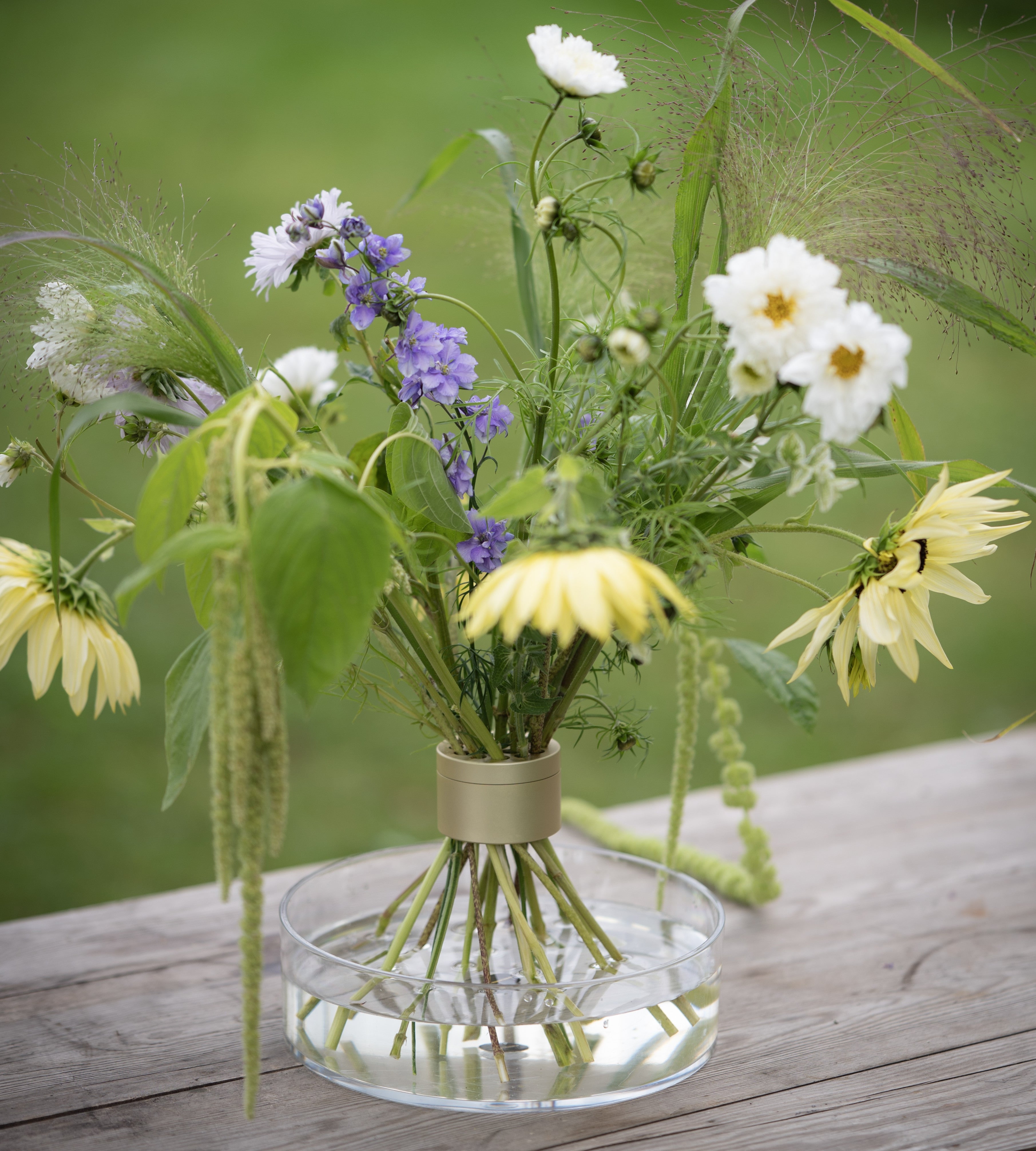 Bouquet of wildflowers inside a Hanataba Bouquet Twister champagne gold standing on a clear vase on a wooden surface with a blurred green background