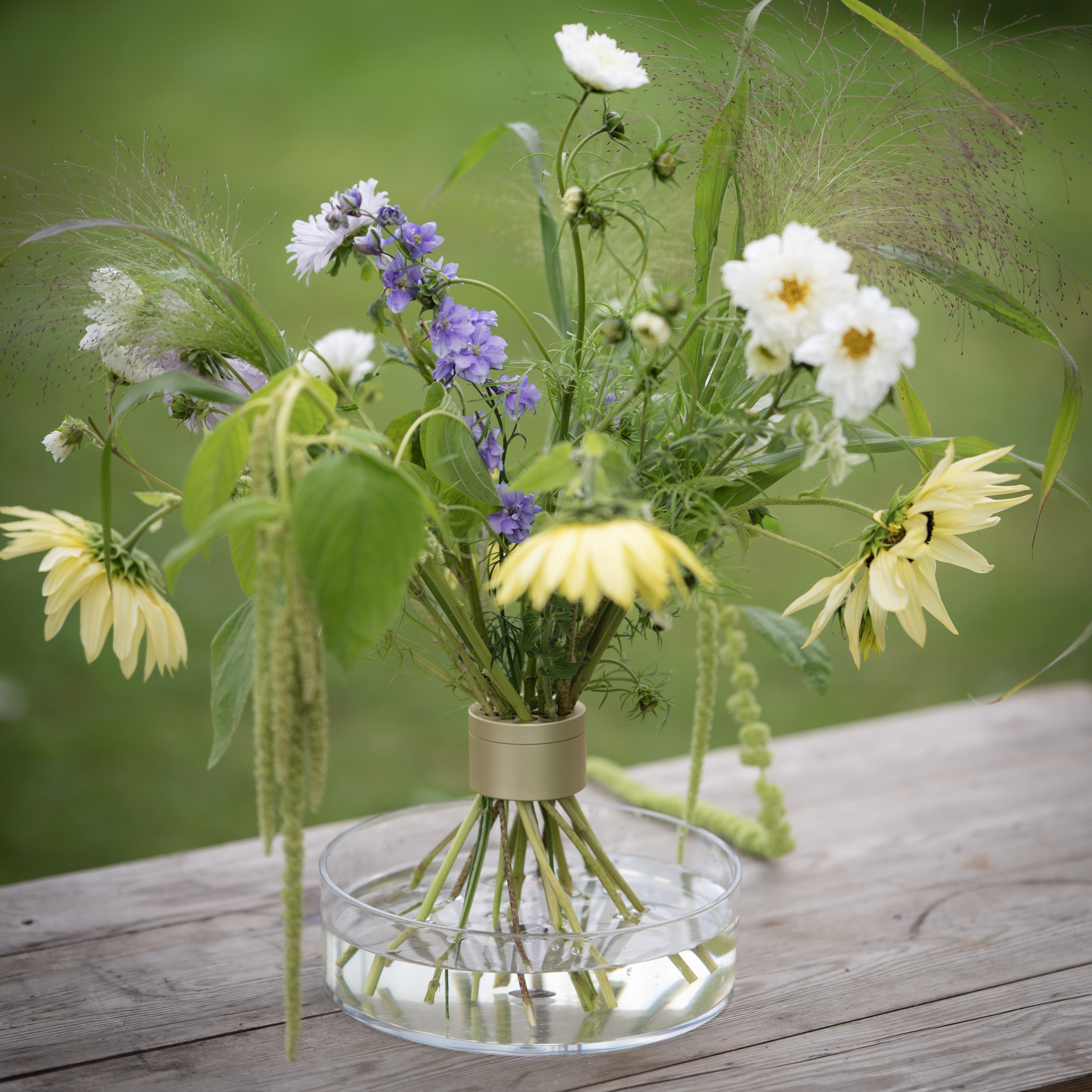 Bouquet of wildflowers inside a Hanataba Bouquet Twister champagne gold standing on a clear vase on a wooden surface with a blurred green background