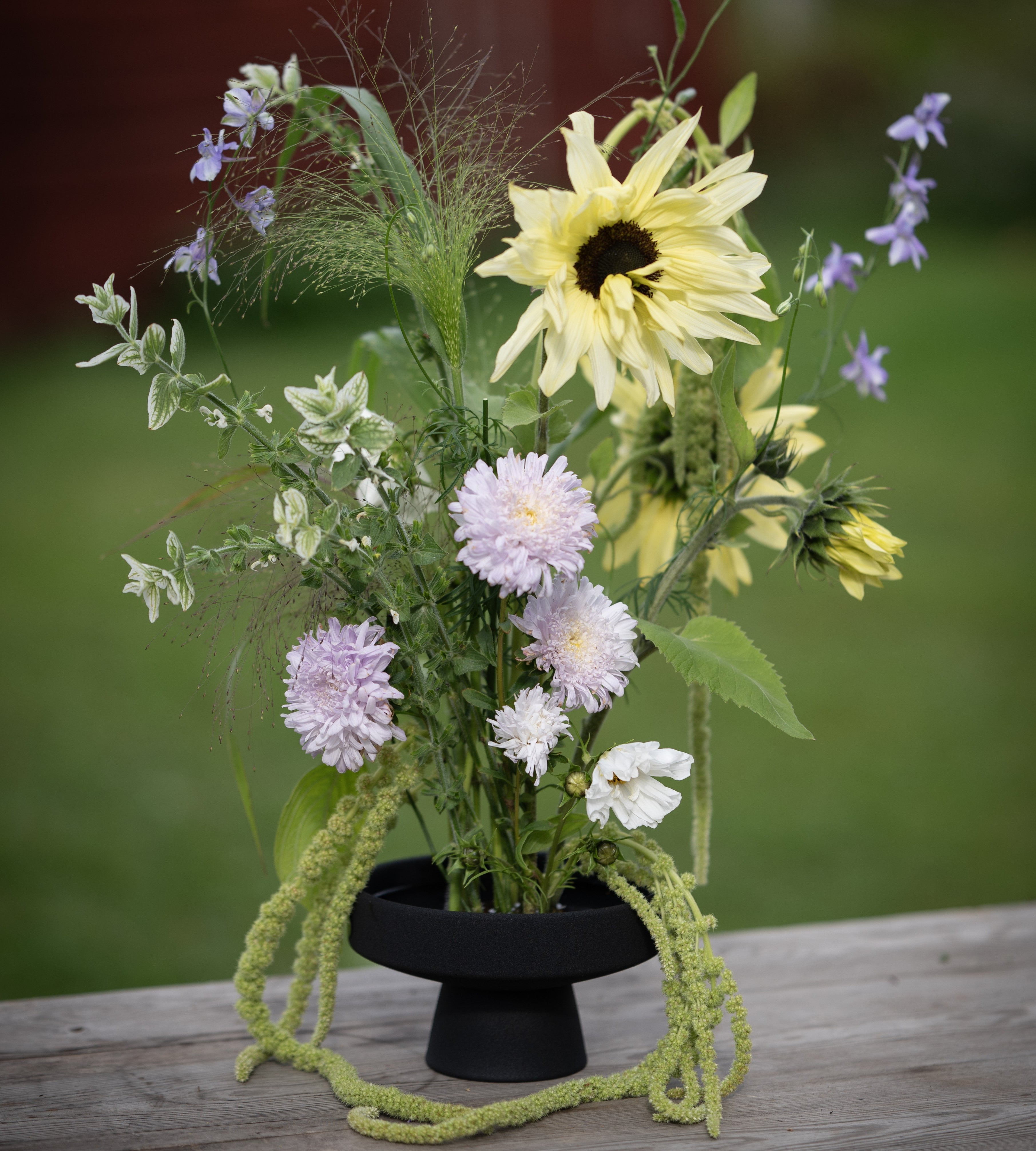 Floral arrangement with sunflowers, asters, and other flowers on a black ceramic ikebana pedestal vase against a blurred natural background.