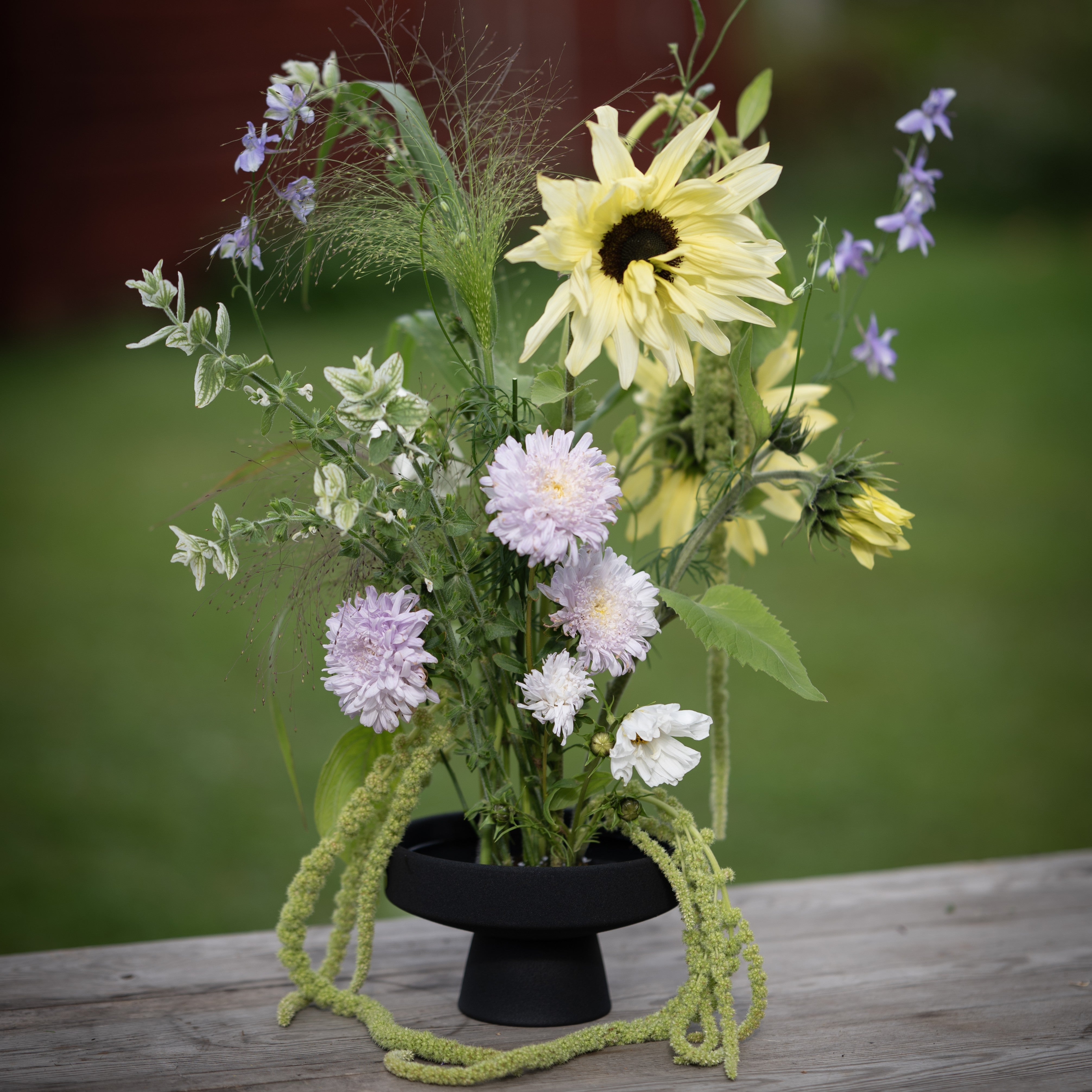 Floral arrangement with sunflowers, asters, and other flowers on a black ceramic ikebana pedestal vase against a blurred natural background.