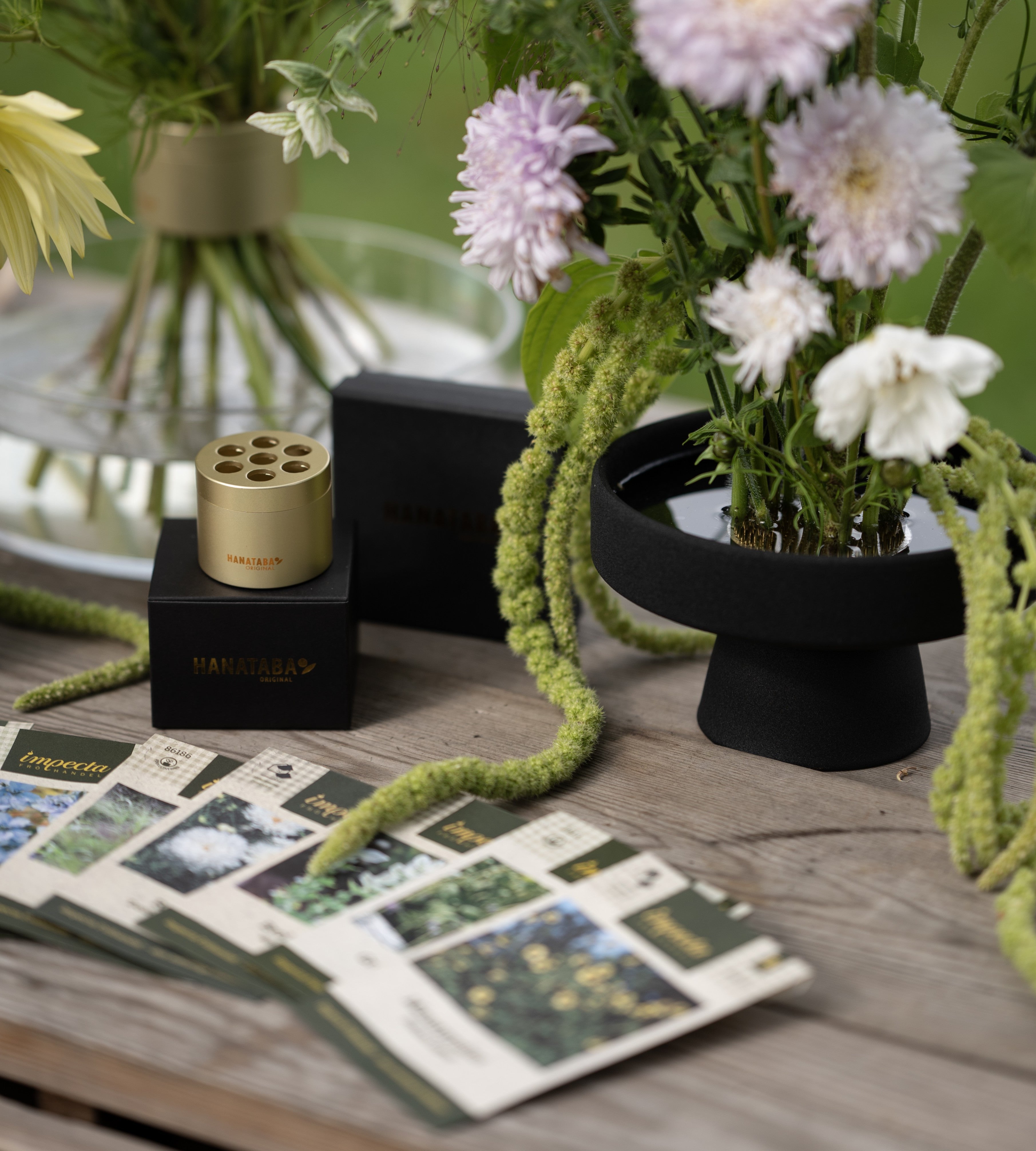 Floral arrangement with a black ceramic kenzan ikebana pedestal vase on a wooden surface, surrounded by seed packets and a small container.