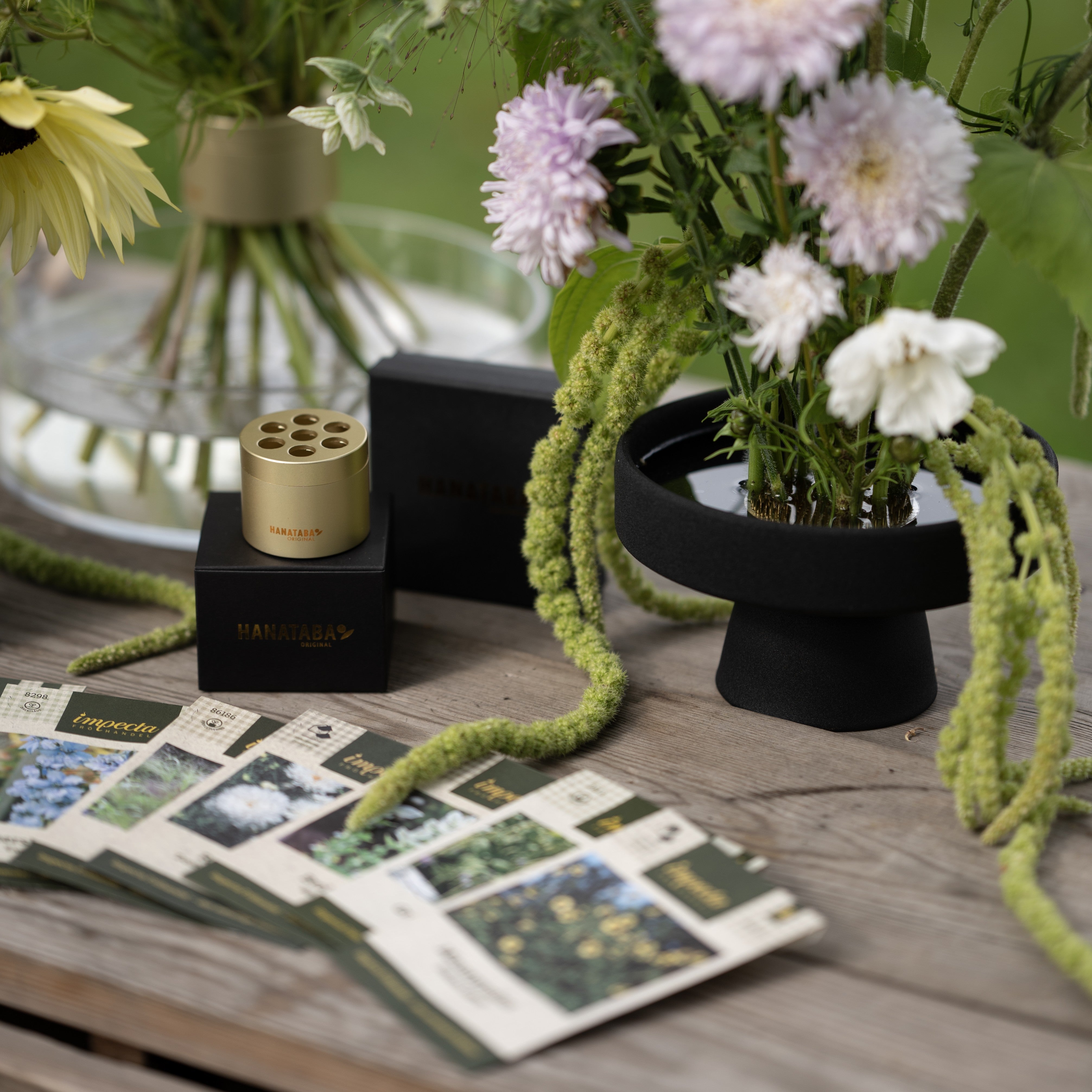 Floral arrangement with a black ceramic kenzan ikebana pedestal vase on a wooden surface, surrounded by seed packets and a small container.