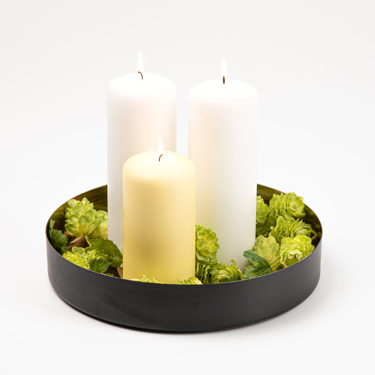 Three pillar candles in white and pale yellow arranged in a round black and gold metal tray with green hop cones, photographed on a clean white background.