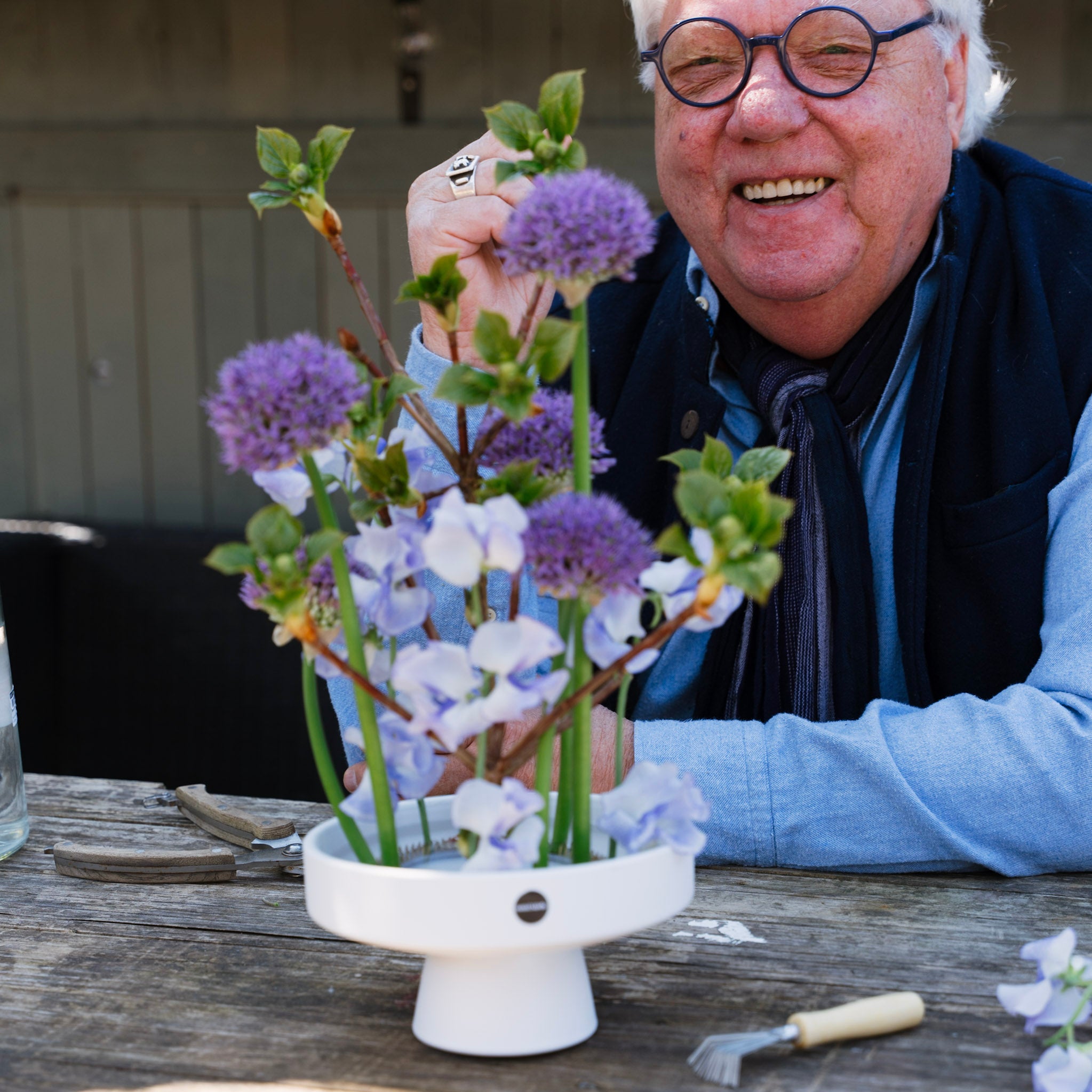 Man arranging flowers in a white vase on a wooden table outdoors.