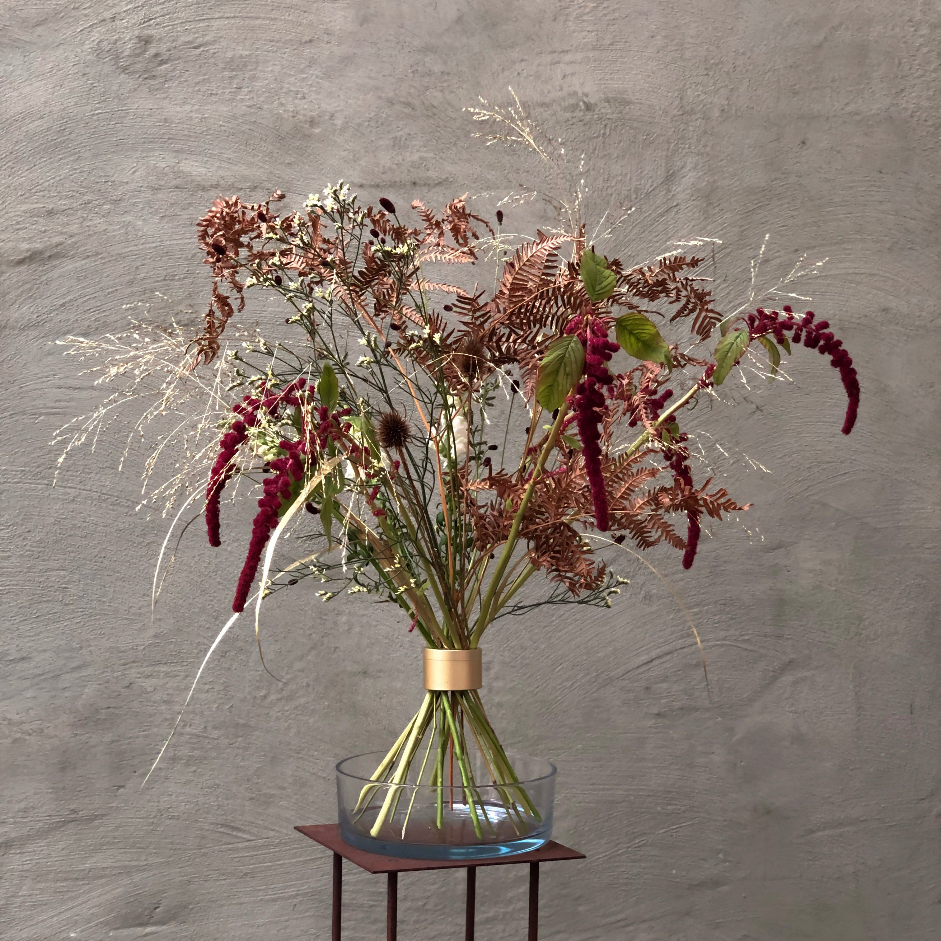 A dried floral arrangement in a clear glass bowl vase with a gold hanataba bouquet flower twister, displayed on a small dark wooden stand against a textured gray wall.