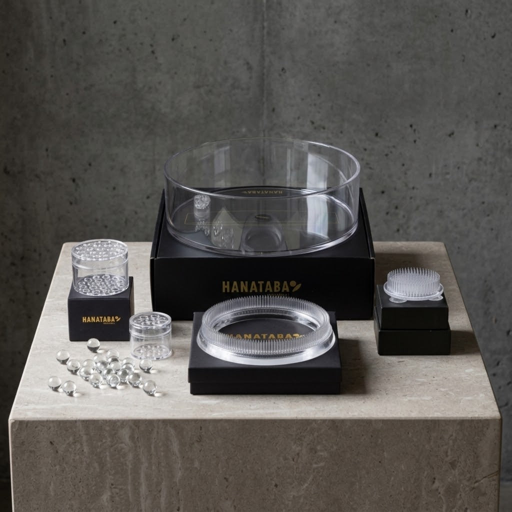 Hanataba clear glass and acrylic floral arrangement set arranged on a stone pedestal against a gray concrete wall, including a large clear bowl on a black base, smaller clear containers, a clear lid, and a white scrubber brush.