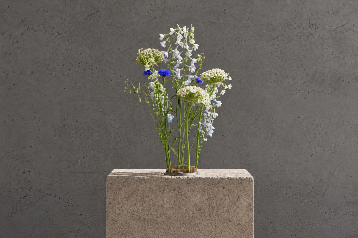 A small arrangement of white and light blue flowers with a few blue blossoms in a Hanataba Kenzan flower frog ring 120 mm, placed on a square concrete pedestal against a textured gray wall.