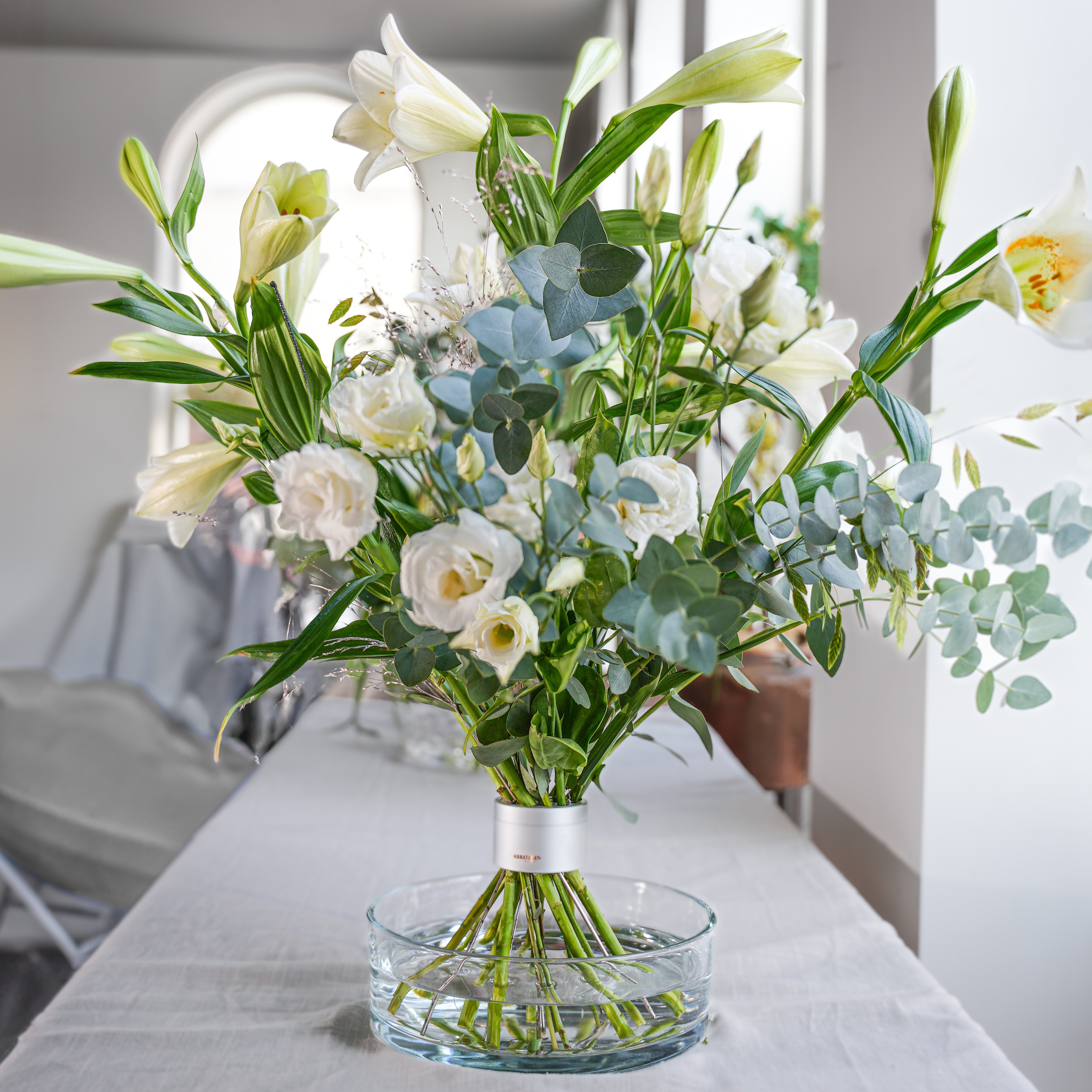 Hanataba pearly silver bouquet of flowers in a clear shallow glasvase on a table with a blurred indoor background