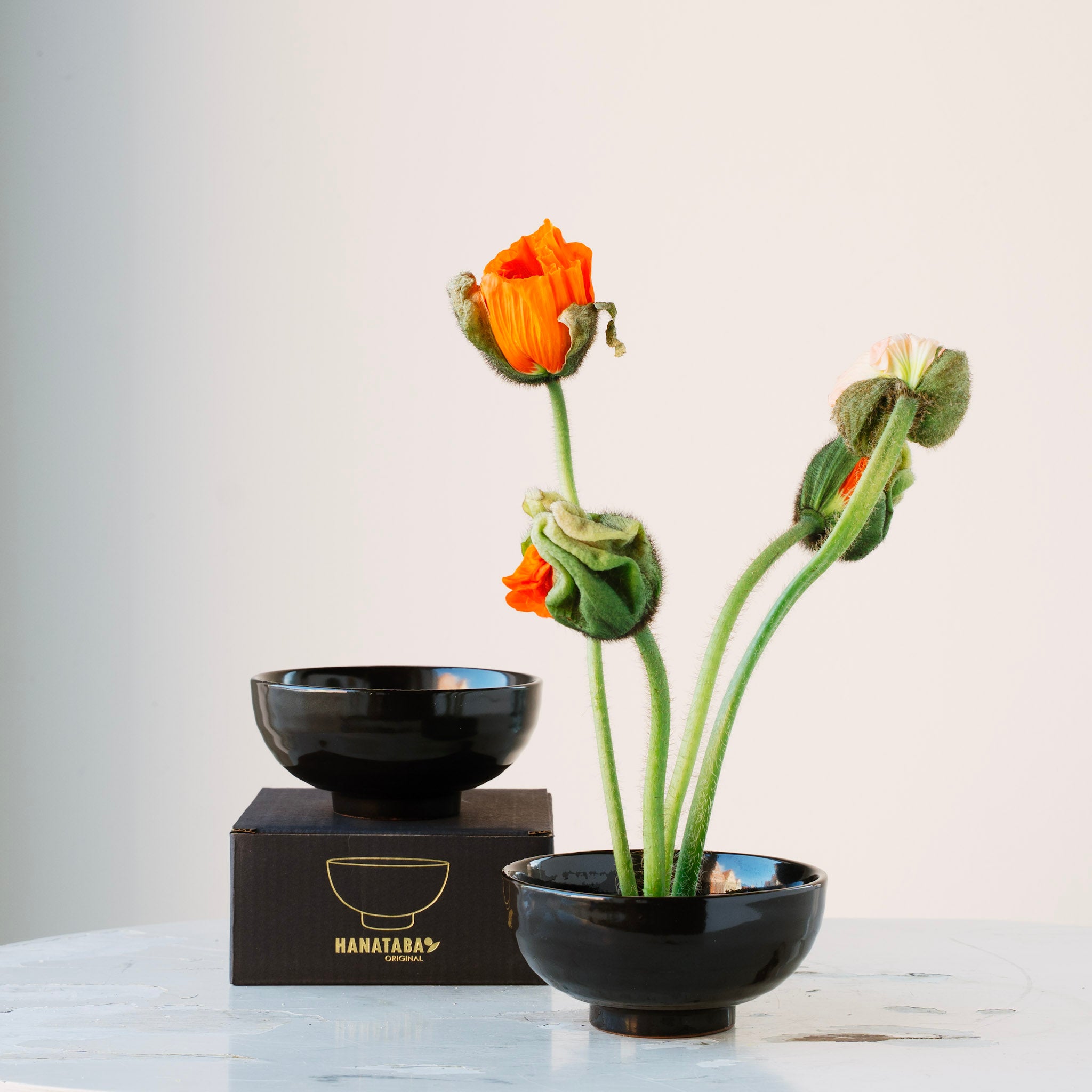 Flower arrangement in a black ceramic kenzan 'kobashi' bowl with a box labeled 'HANATABA' on a light background