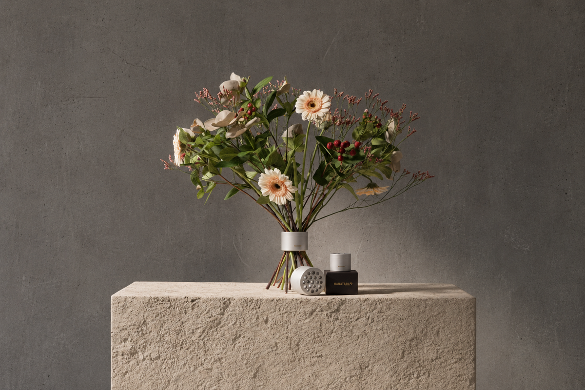 A winter flower bouquet arrangement in a Hanataba Bouquet Flower Twister Pearly silver, displayed on a textured stone pedestal against a dark gray concrete wall. A small dark bottle and a round patterned object sit beside the vase.