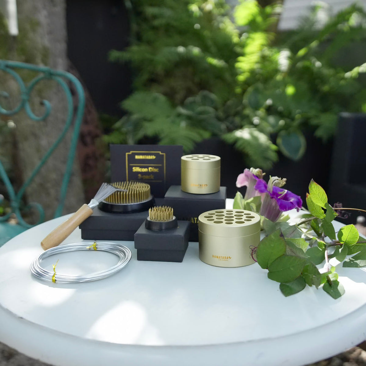 A grooming kit with hanataba bouquet Twister Champagne Gold, black paper boxes, a wooden kenzan 7 cm and 3,4 cm tool, and silver wire coiled on a white table outdoors, styled with green leaves and purple flowers.