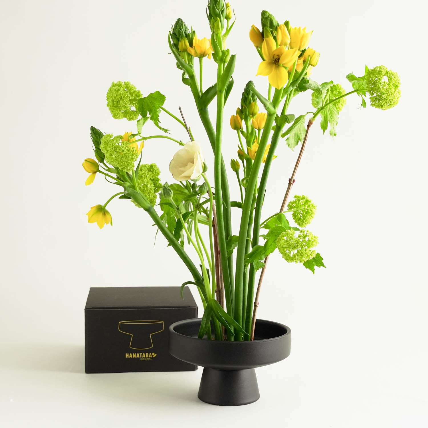 A black, pedestal-style vase with a Japanese ceramic touch holds an arrangement of yellow and white flowers alongside lush green foliage. Behind the vase is a black packaging box with the hanataba logo in gold, all set against a plain white background.