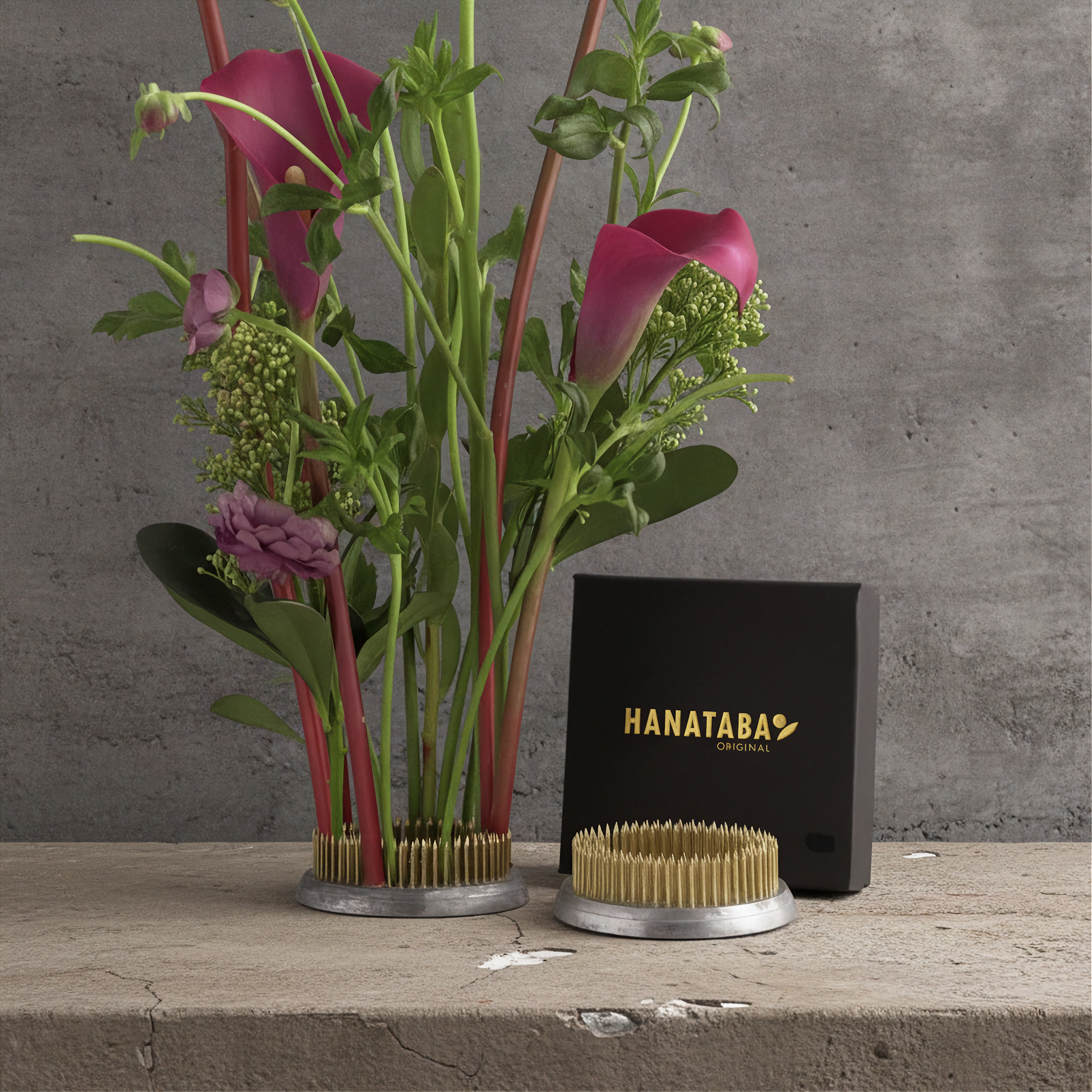 A floral arrangement with pink calla lilies and green foliage in a kenzan flower frog pin holder, shown beside a black box labeled HANATABA on a concrete surface against a gray wall.