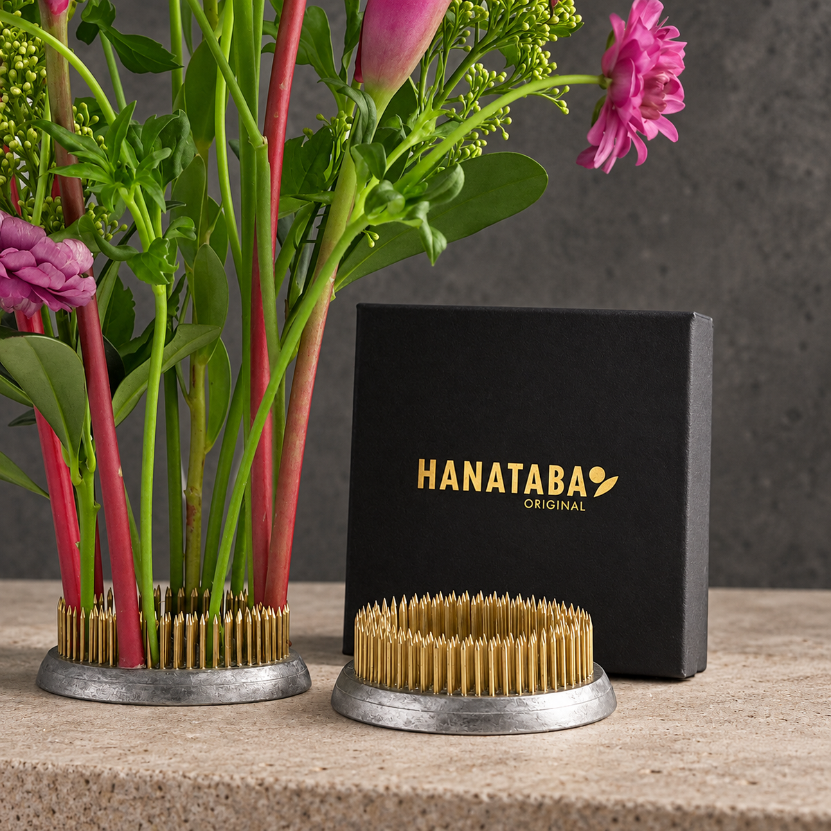 A pair of Hanataba Original kenzan ring 70 mm with a gold-colored sharpening spike pattern and silver metal bases, displayed next to a black branded gift box on a stone surface with a floral arrangement in the background.