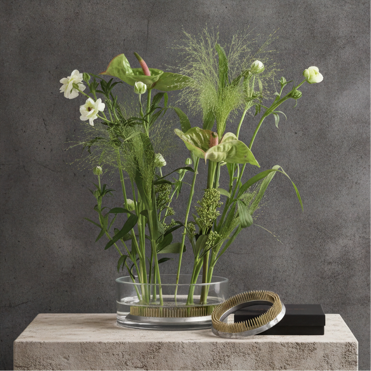 A clear glass vase filled with green and white botanical stems, placed on a light stone pedestal against a textured gray wall. A Hanataba Original Kenzan ring 120 mm and a small black box rest beside the vase.