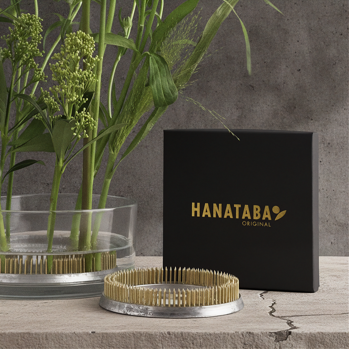 A Hanataba Original product display featuring a black branded box, a round metal tray with light wood bristles, and a glass planter with green stems against a textured gray wall.
