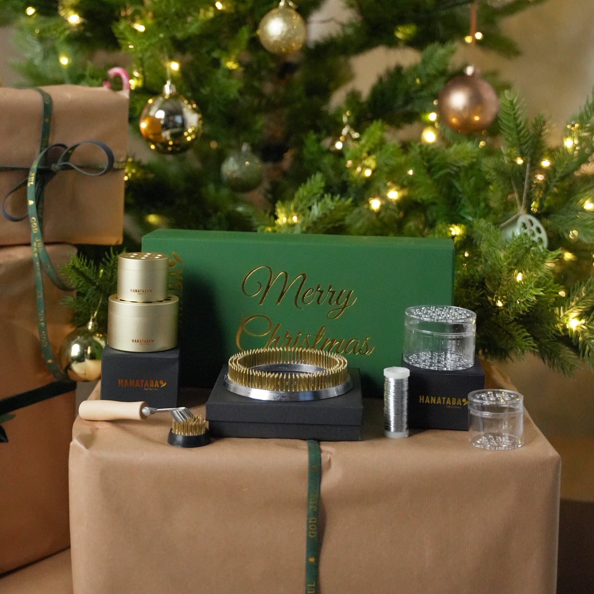 A Christmas gift set in a green box with "Merry Christmas" in gold, featuring flower arranging essentials such as a 120mm kenzan ring, a 34mm flower frog and four Hanataba flower twisters. The products are displayed elegantly on a light surface.