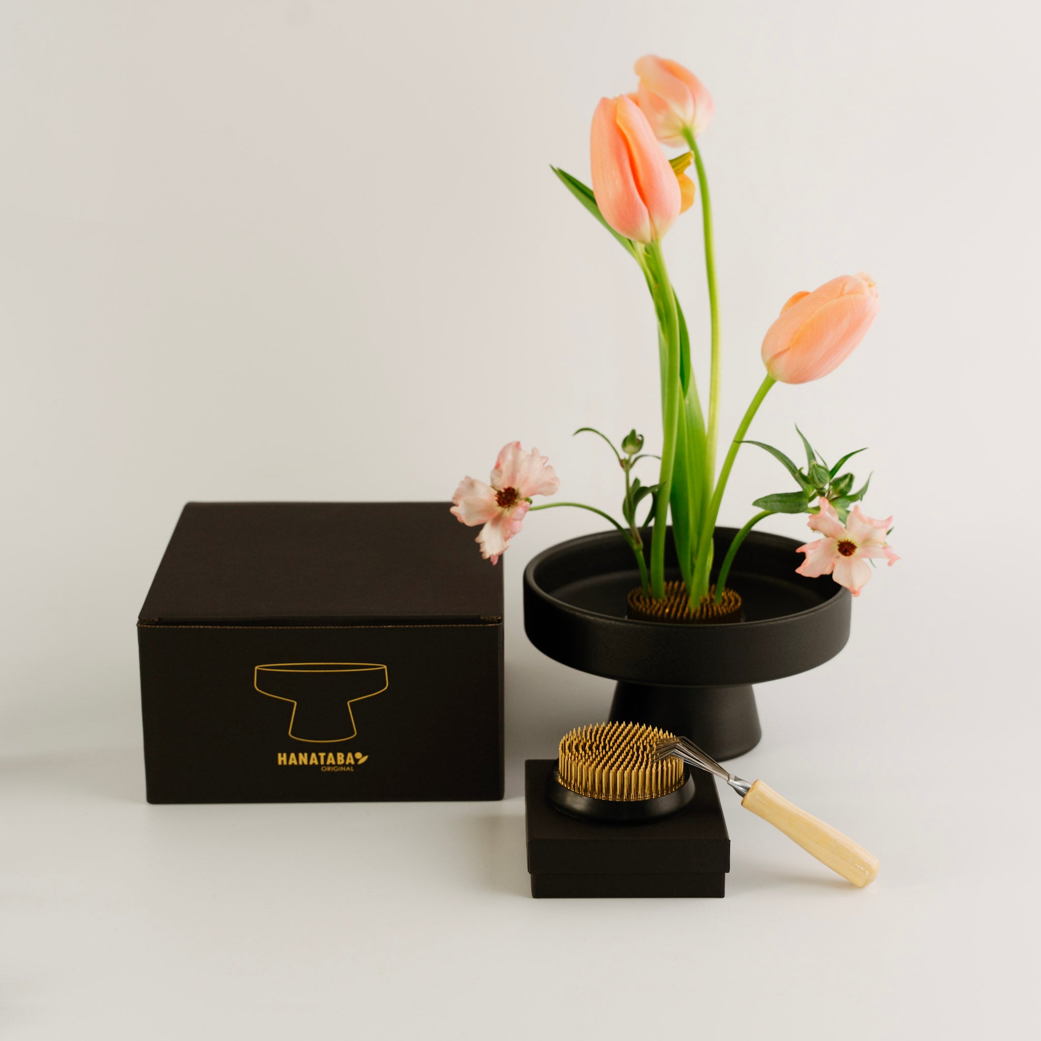 Black box with gold logo 'HANATABA', black stand with flowers, and brush on a white background