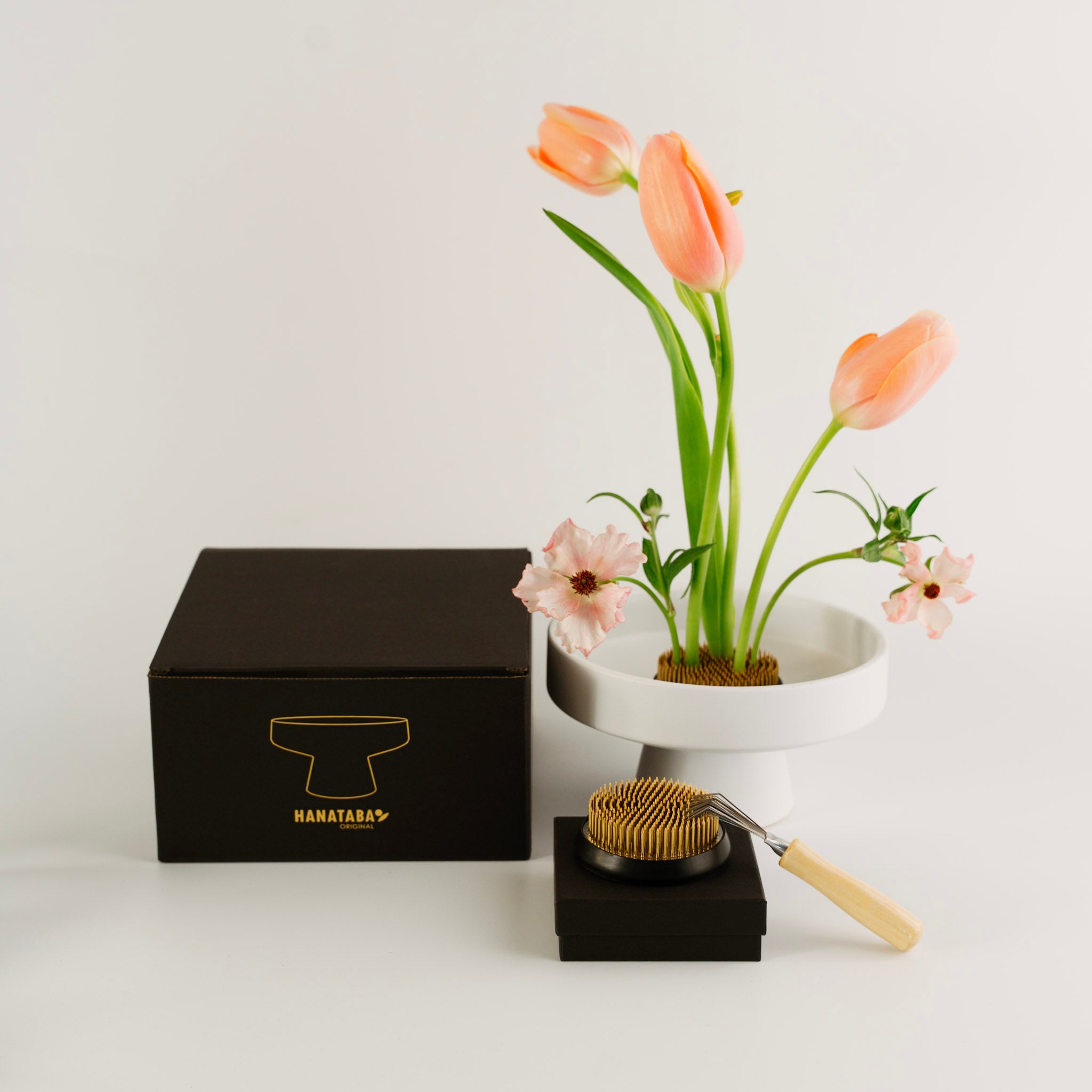 Black box with gold 'HANATABA' logo, white vase with pink tulips, and wooden tool on a light gray background