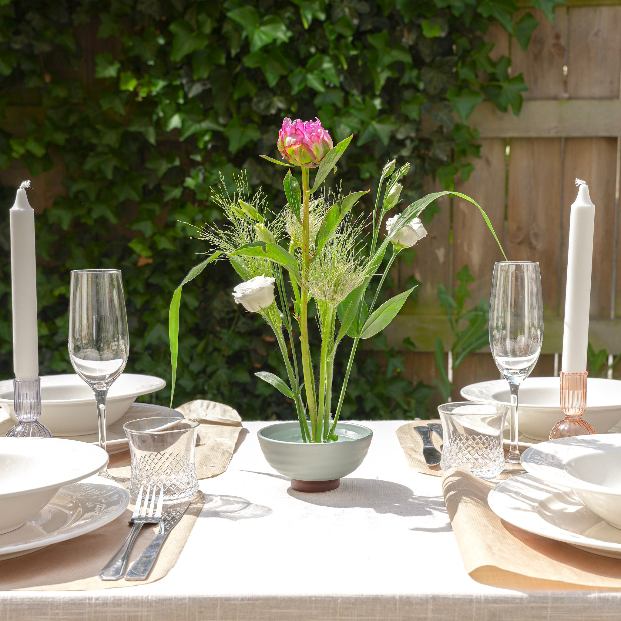 Outdoor table setting with floral centerpiece inside a ceramic kenzan 'kobachi' bowl, candles, and glasses.