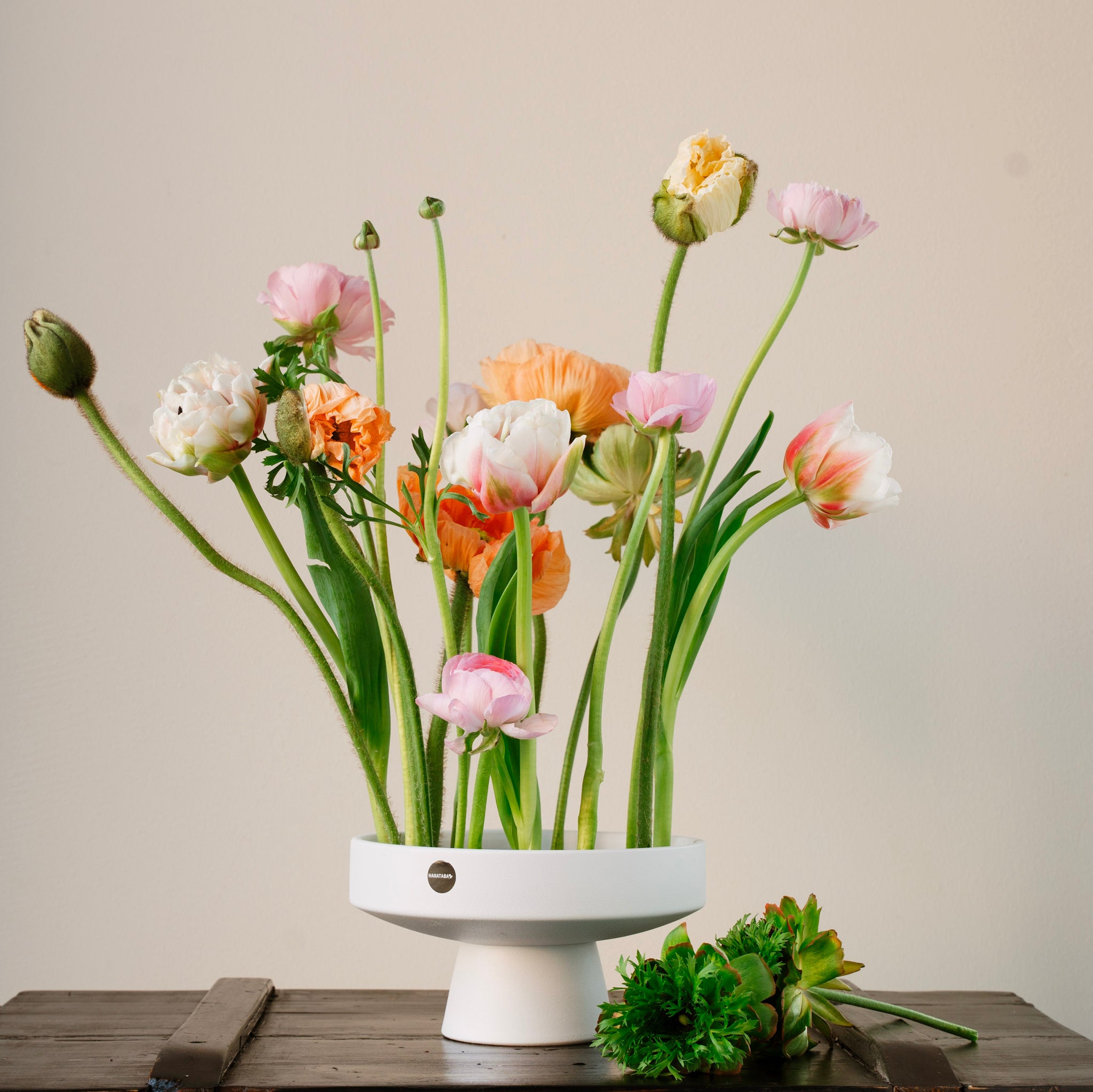 Floral arrangement in a white hanataba ceramic ikebana vase on a wooden surface with a neutral background