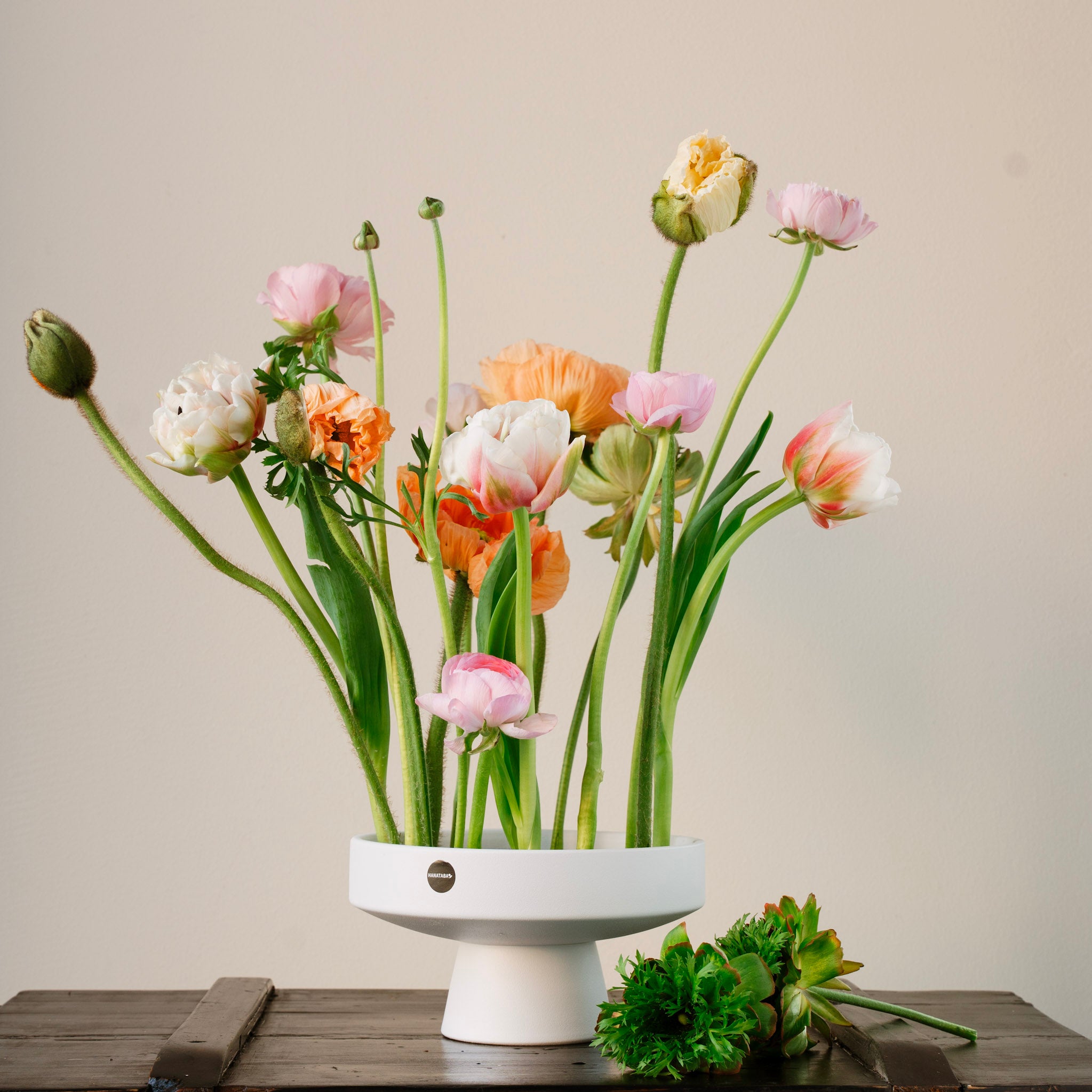 Floral arrangement in a white hanataba ceramic ikebana vase on a wooden surface with a neutral background