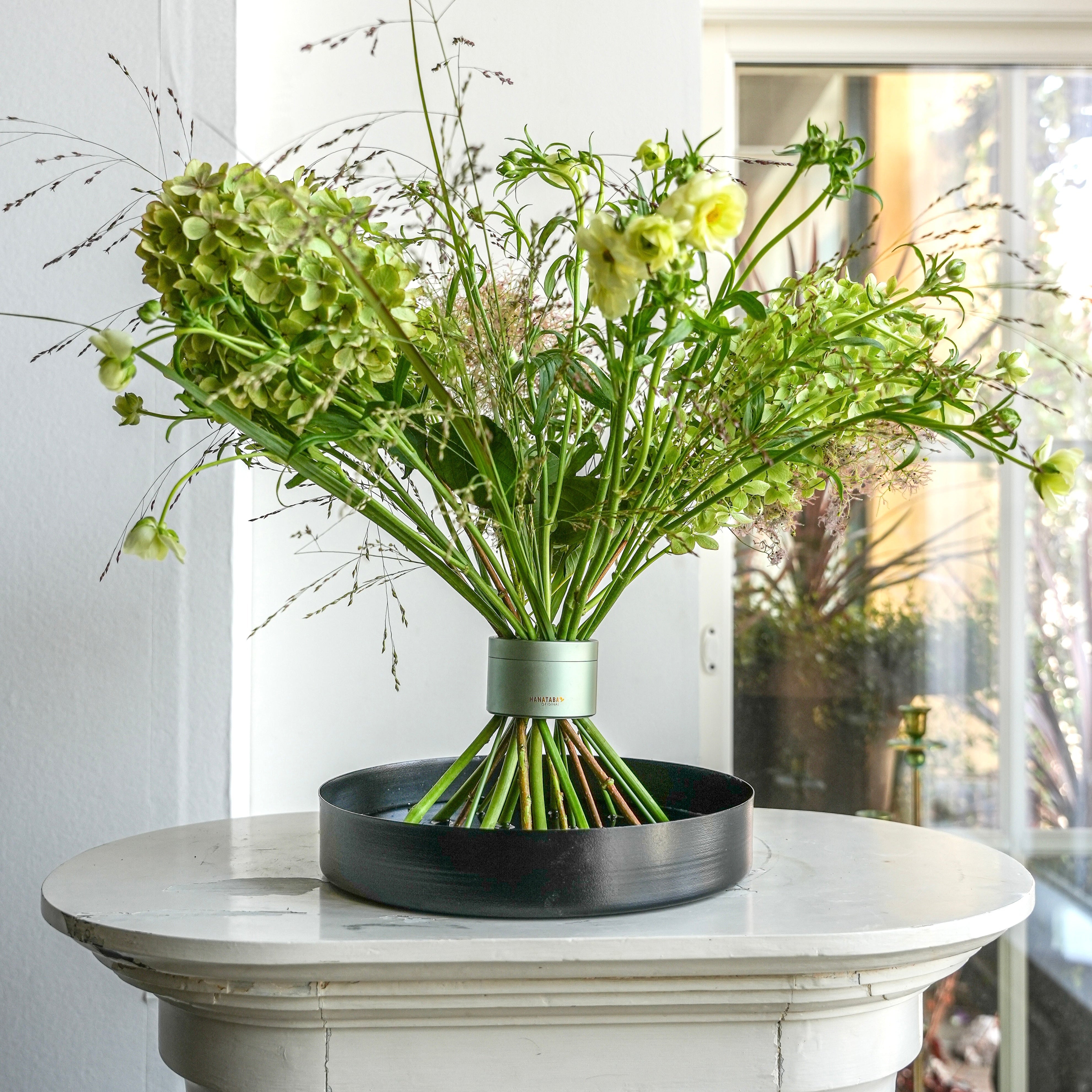 Professional bouquet with beautiful volume in Hanataba Matcha Green flower twister, displayed on a black floral tray atop a white pedestal.