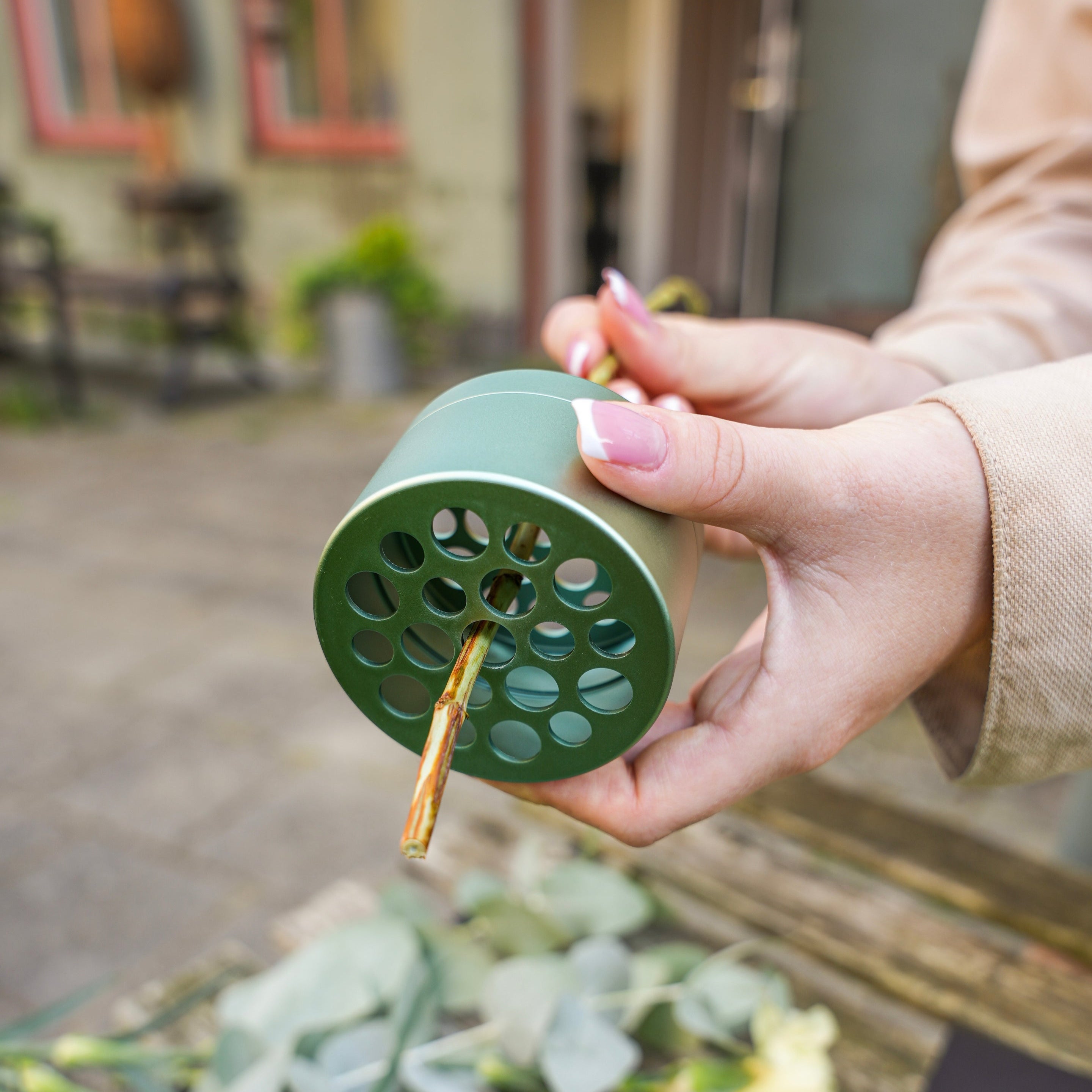 A person holding an Hanataba Ivy Green floral frog with a flower stem inserted through one of its holes. Eucalyptus leaves and other greenery are scattered on the surface below. The background shows an outdoor setting.
