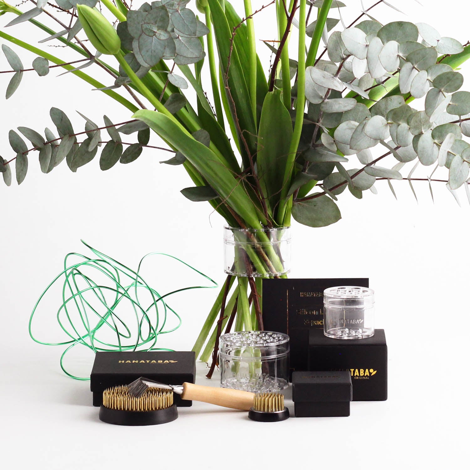 The Home Florist’s Starter Kit – Crystal Clear by Hanataba features an elegant bouquet in a vase with eucalyptus leaves and tulips, surrounded by twisted green bonsai aluminum wire. The kit includes transparent Hanataba Flower Twisters, a 70 mm flower frog, a 34 mm flower frog, a kenzan brush, and black Hanataba packaging boxes, all beautifully arranged on a white surface.
