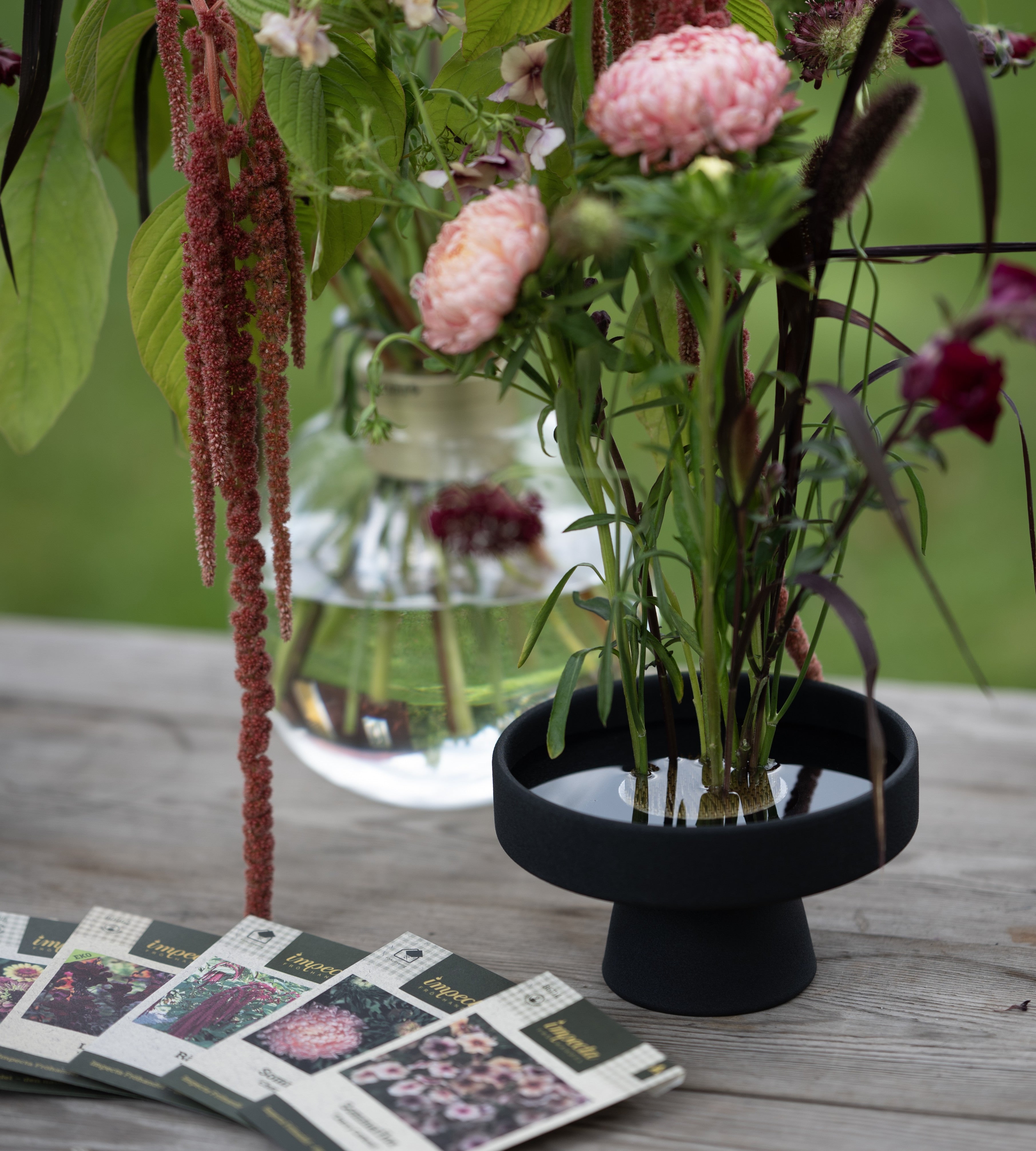 Floral arrangement in a black hanataba ceramic ikebana vase on a wooden surface with seed packets from impecta fröhandel.