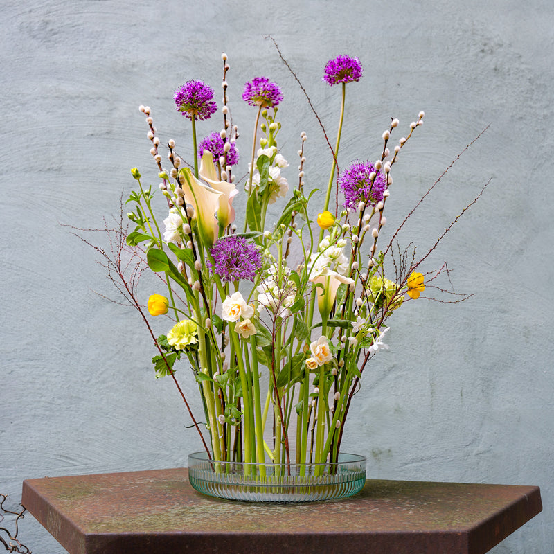 A diverse ikebana arrangement with alliums, calla lilies, and pussy willow, anchored by a 200mm Kenzan Fakir ring, set against a textured grey background.