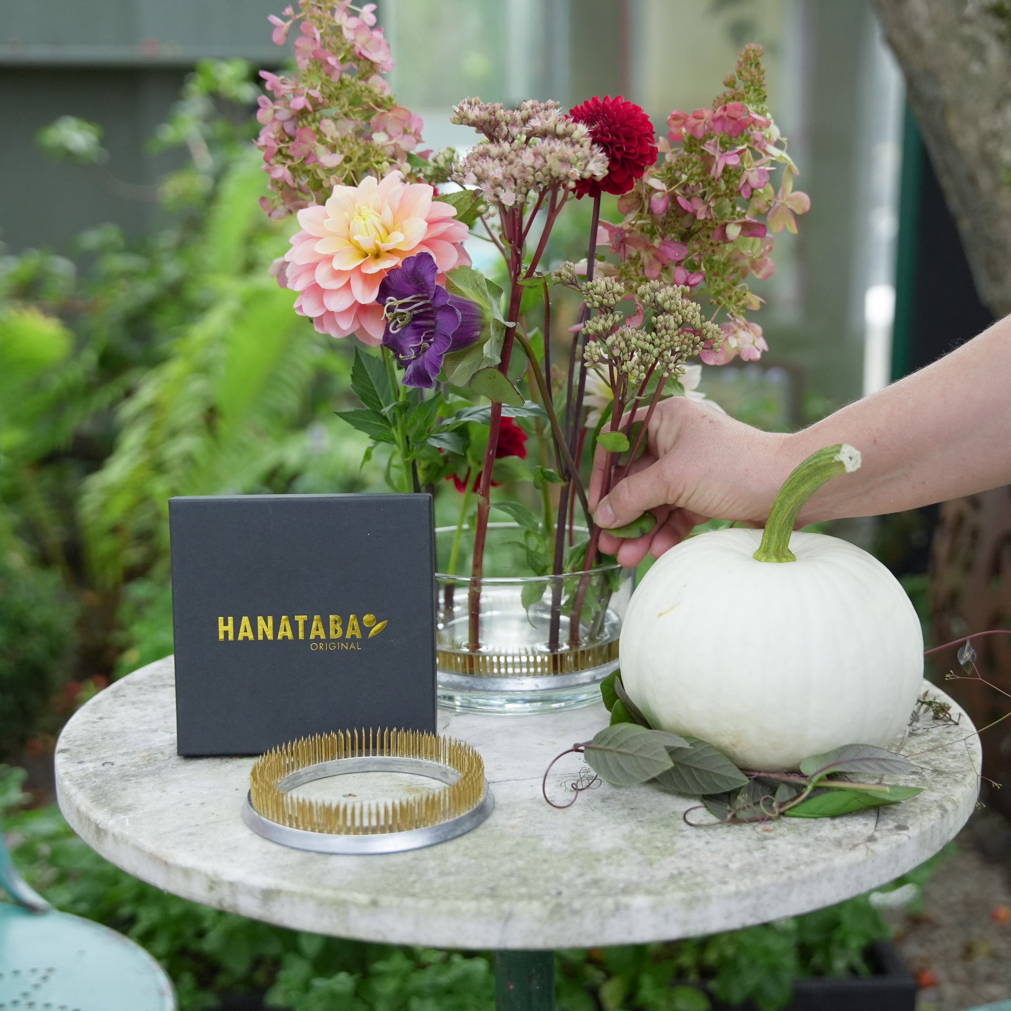 Table with a flower arrangement on a kenzan flower frog ring 120 mm, a white pumpkin, and a 'HANATABA' box in an outdoor setting.