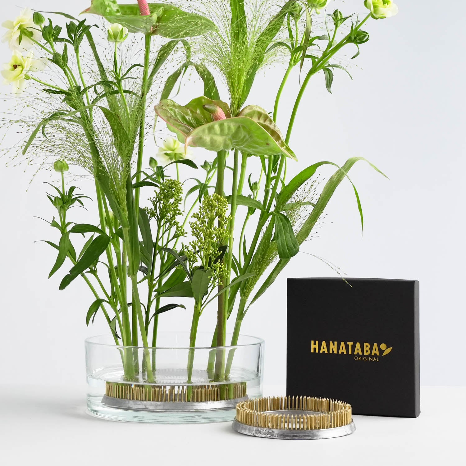 A glass vase with assorted green and pink flowers is displayed next to a black packaging labeled "Hanataba Original" and a metal kenzan flower frog holder. The flowers, held securely by the kenzan, are artfully arranged with varying heights and textures against a light background.