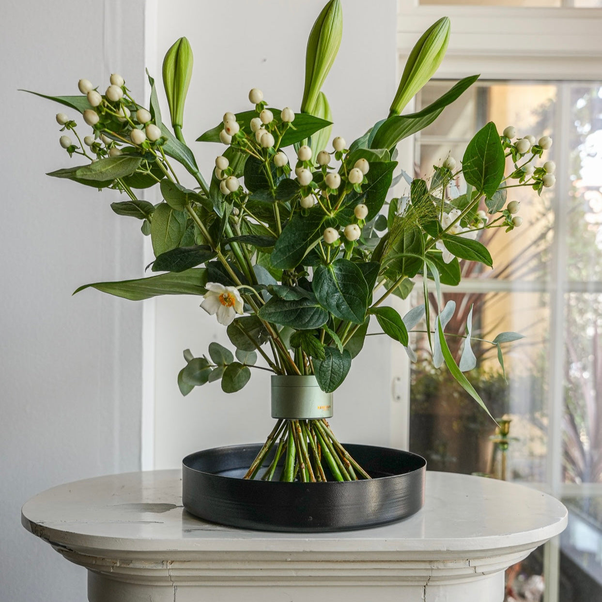 Stunning flower arrangement with lilies displayed on a black floral tray atop a white pedestal in a beautiful white room with paneled windows.