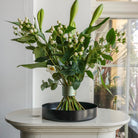 Stunning flower arrangement with lilies displayed on a black floral tray atop a white pedestal in a beautiful white room with paneled windows.